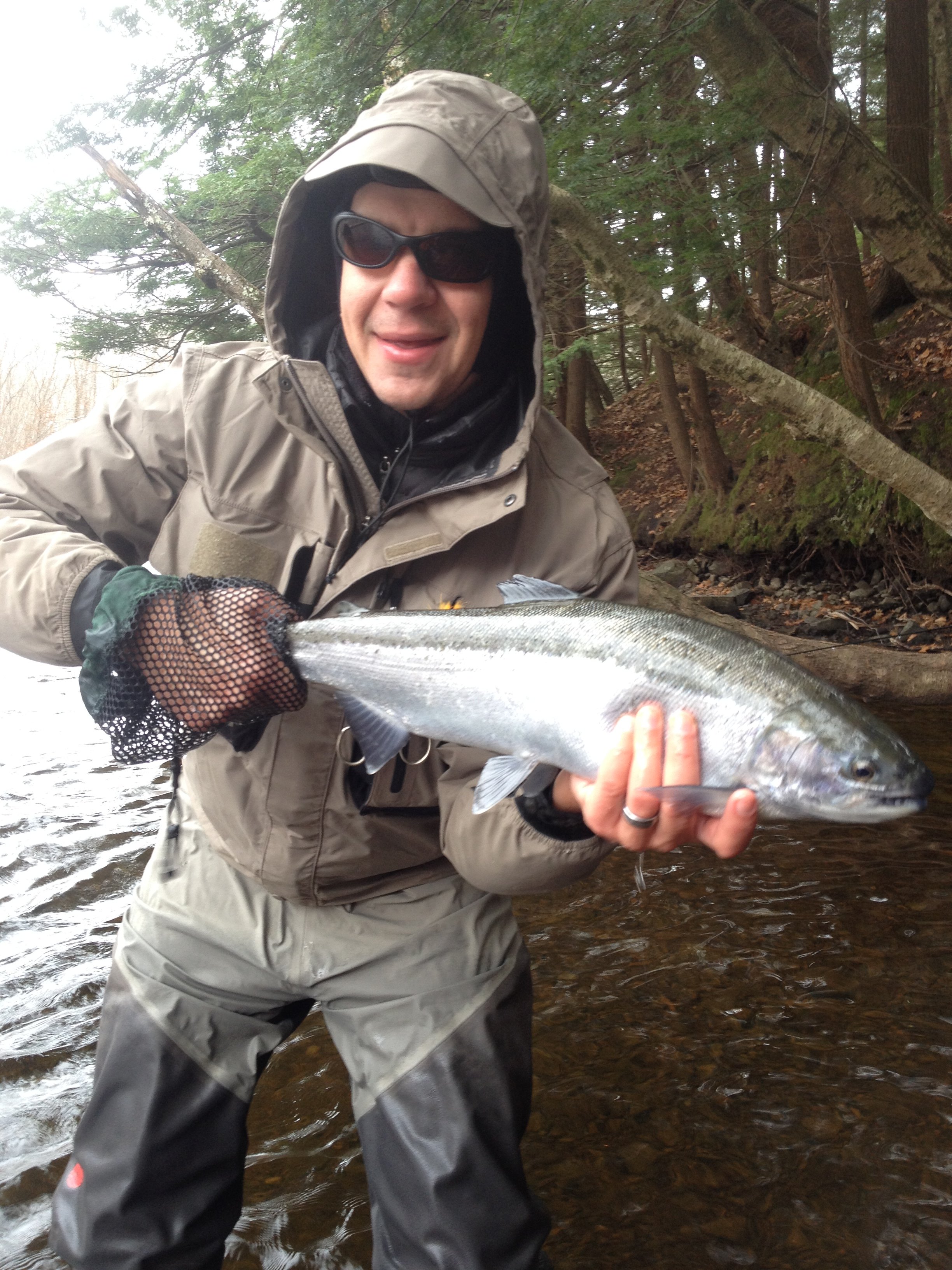 Man dressed in fishing gear holding a large fish in a river with trees in the background.