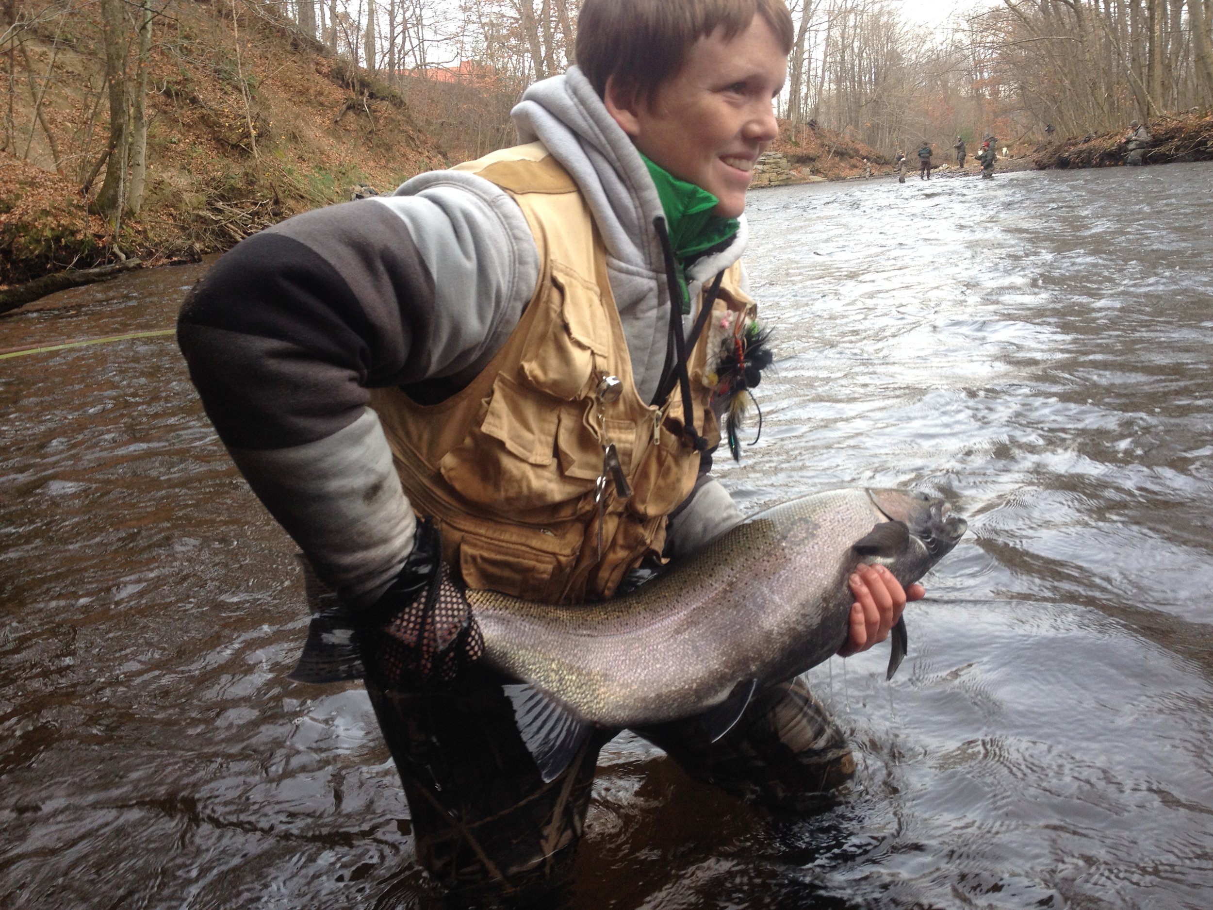 A smiling man in a gray and black hooded jacket with a tan fishing vest is holding a large fish in a river during daytime. trees and other people fishing are visible in the background.