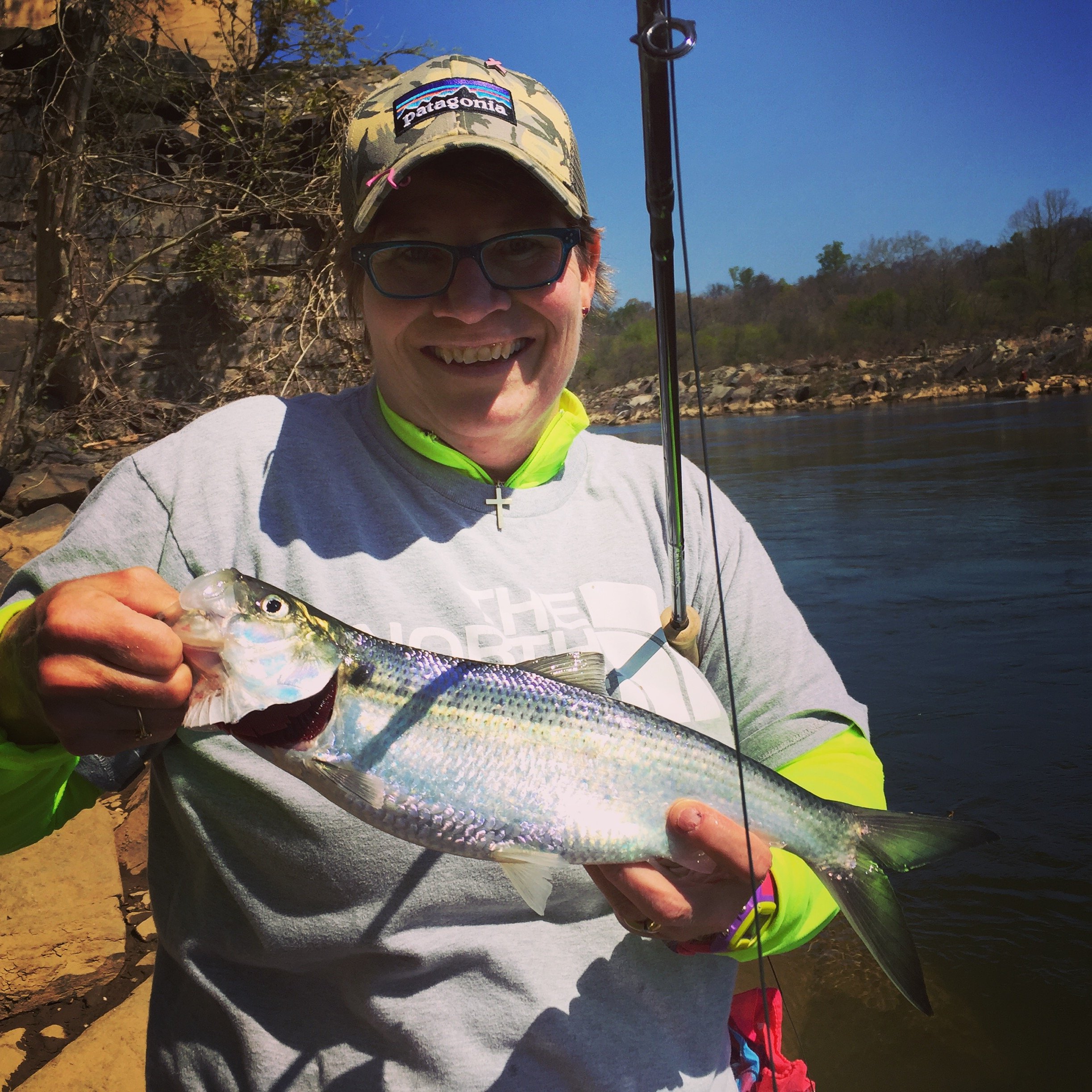 A person smiling and holding a large fish they caught while fishing near a river on a sunny day, wearing glasses, a Patagonia cap, a gray sweatshirt, and a cross necklace.