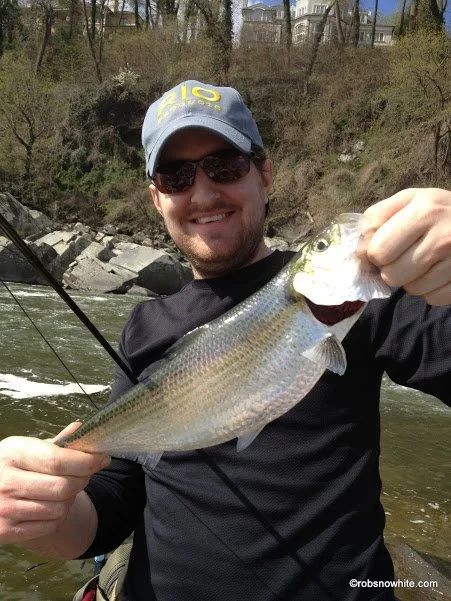 Man smiling, wearing sunglasses and cap, holding a large fish he caught in a river with trees and houses in the background.