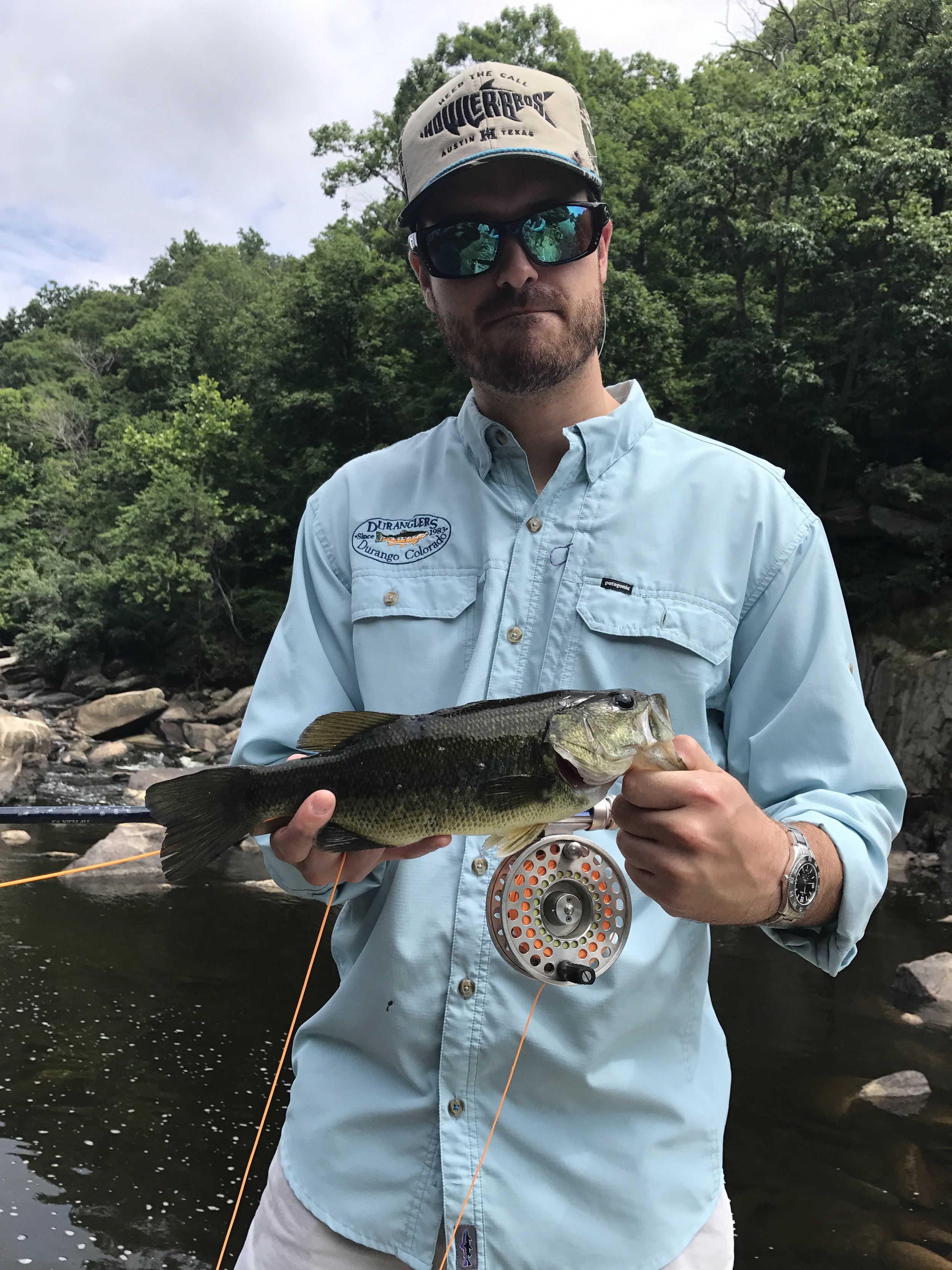 Man holding a largemouth bass caught during a fishing trip, standing in front of a river with trees.