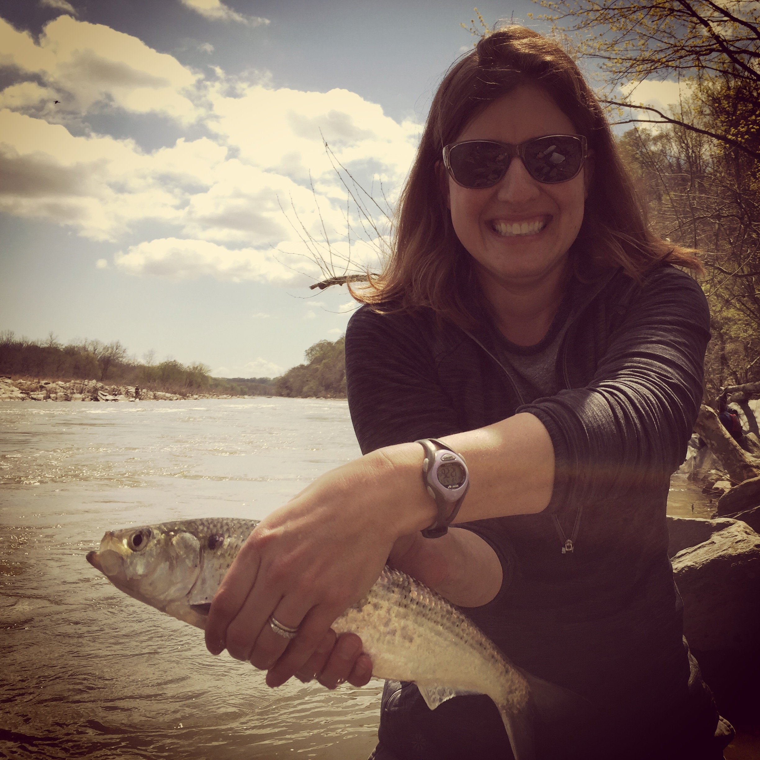A woman wearing sunglasses and a black jacket smiling while holding a fish in her hand at a riverbank during daytime.