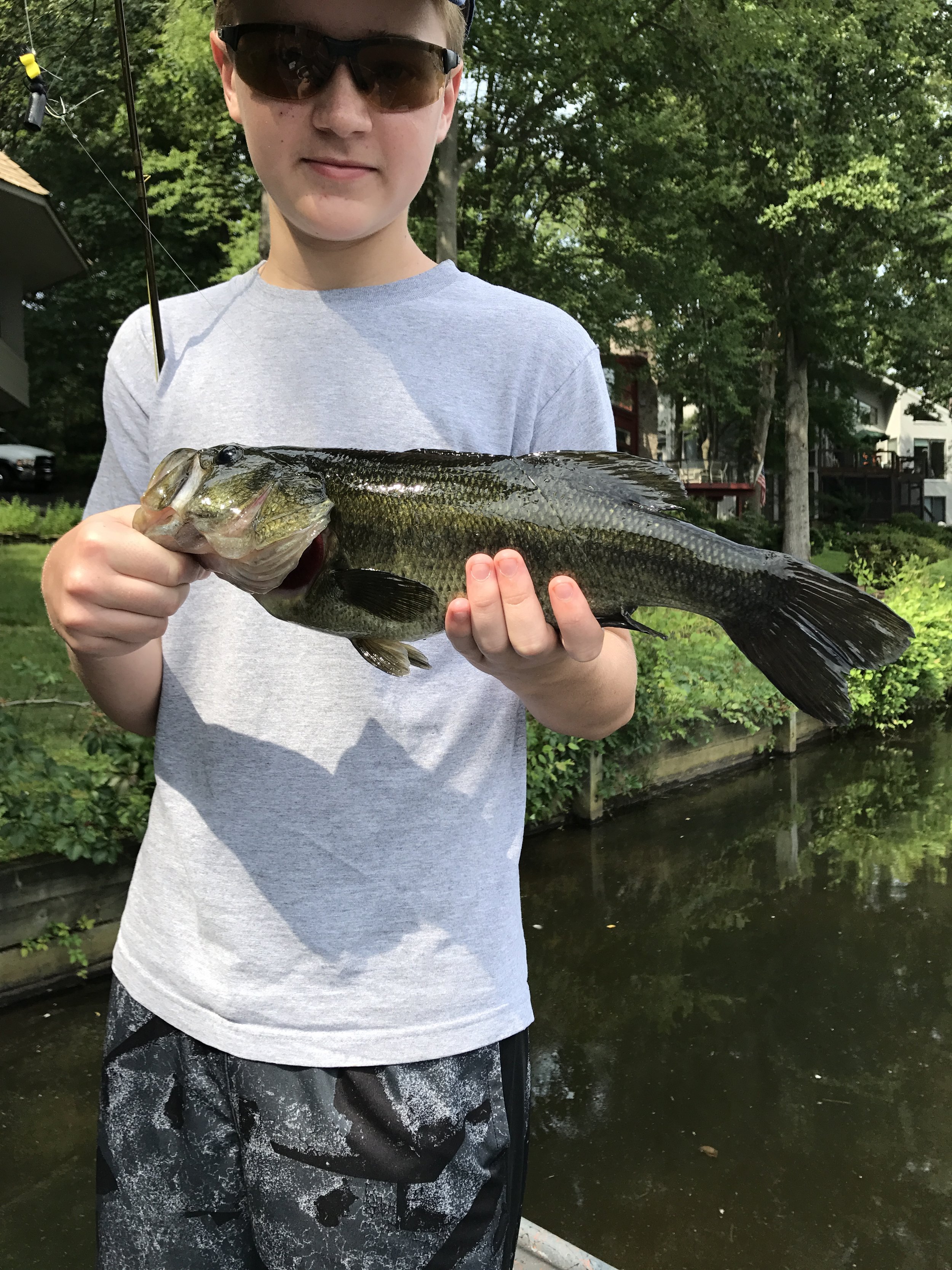 A young boy wearing sunglasses and a gray t-shirt holding a large fish with a greenish-black body and a white belly, standing outdoors near a waterway with trees and houses in the background.