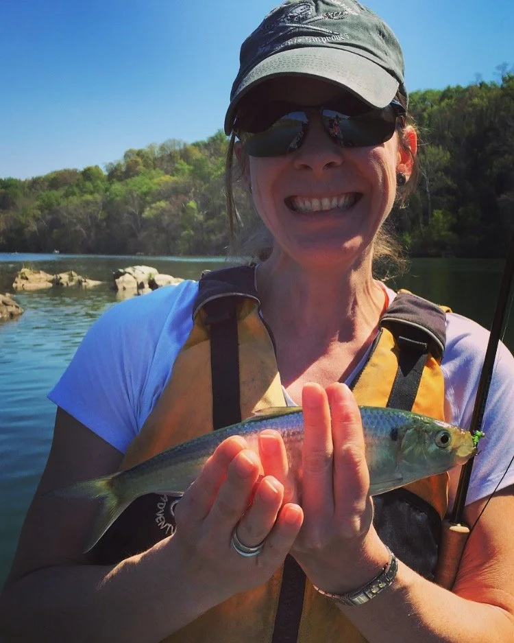 Woman wearing sunglasses, a hat, and a life jacket holding a fish she caught, outdoors on a sunny day with a body of water and trees in the background.