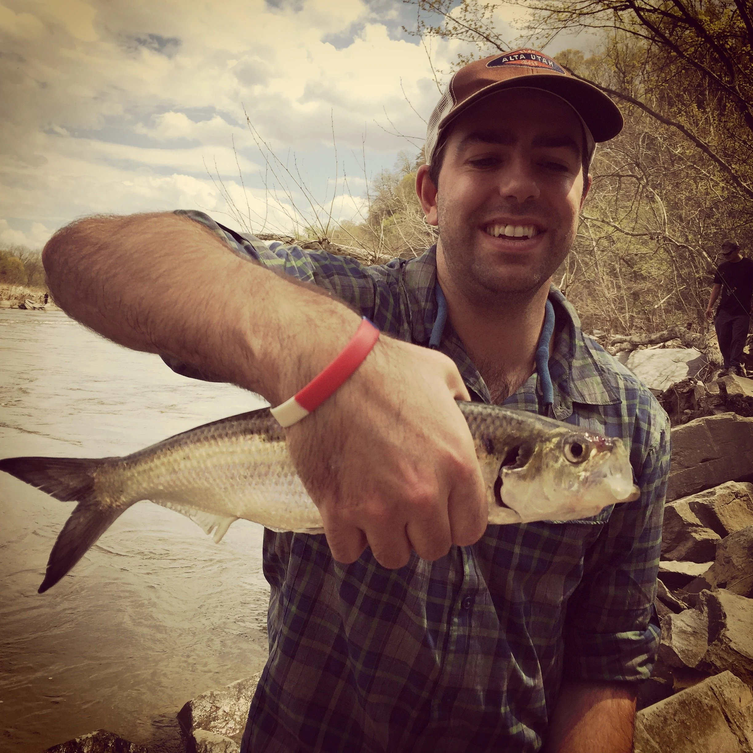 A man smiling and holding a fish near a riverbank with rocks and trees in the background.