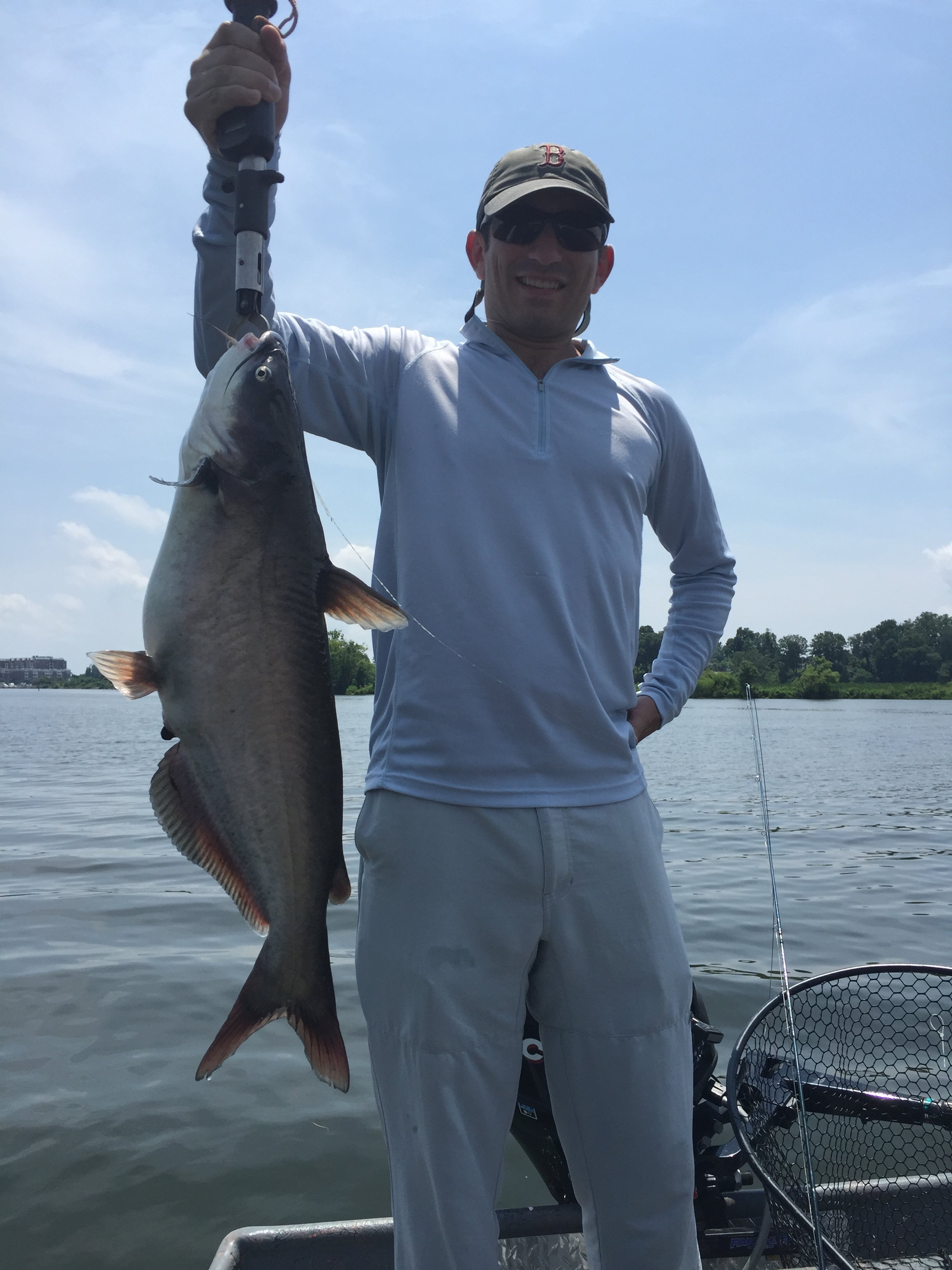 Man in sunglasses and hat holding a large fish over water on a boat with a fishing net nearby.