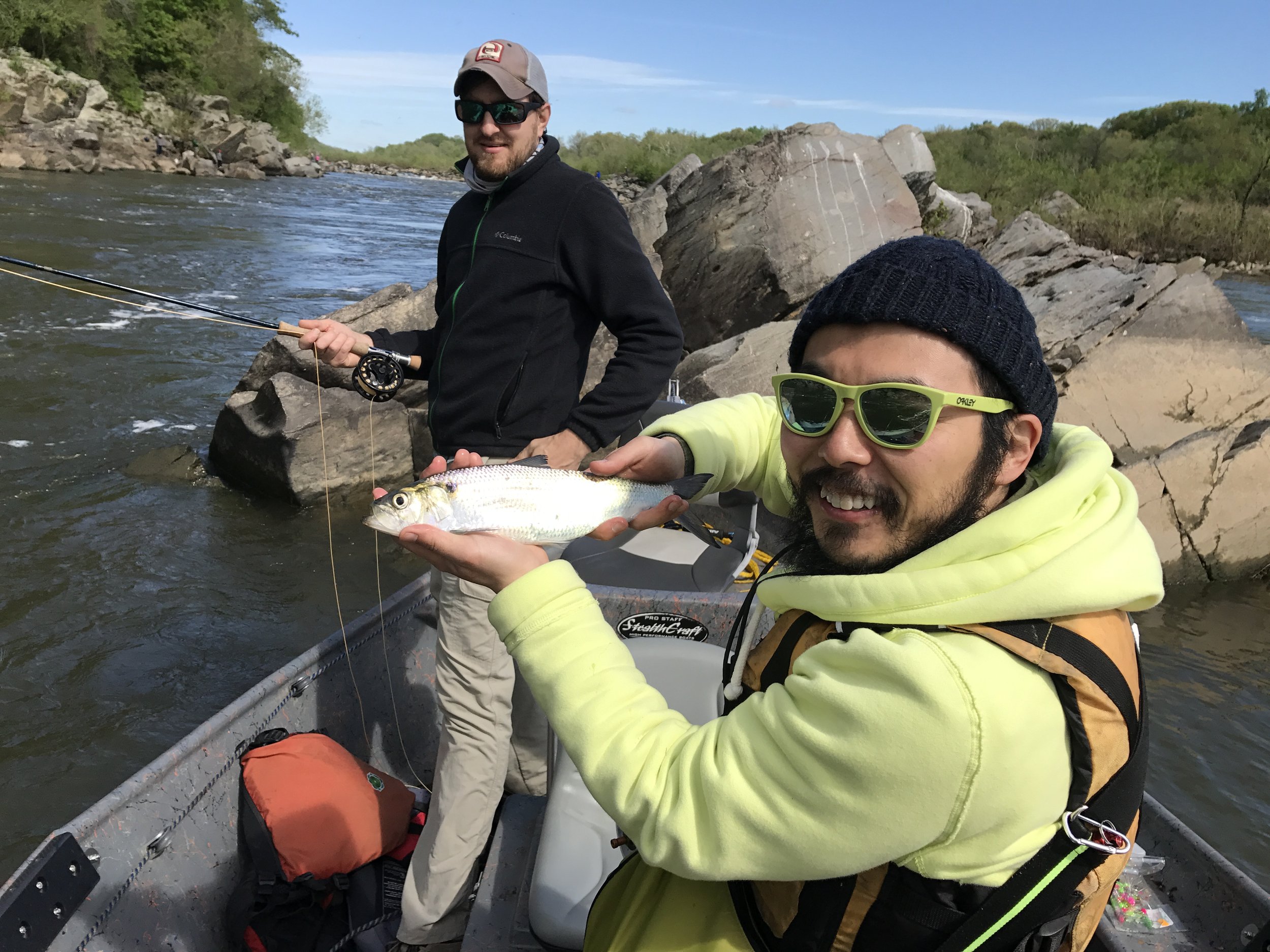 Two men fishing in a river, one holding a fish and smiling at the camera, the other standing in the water with a fishing rod, rocks, and greenery in the background.
