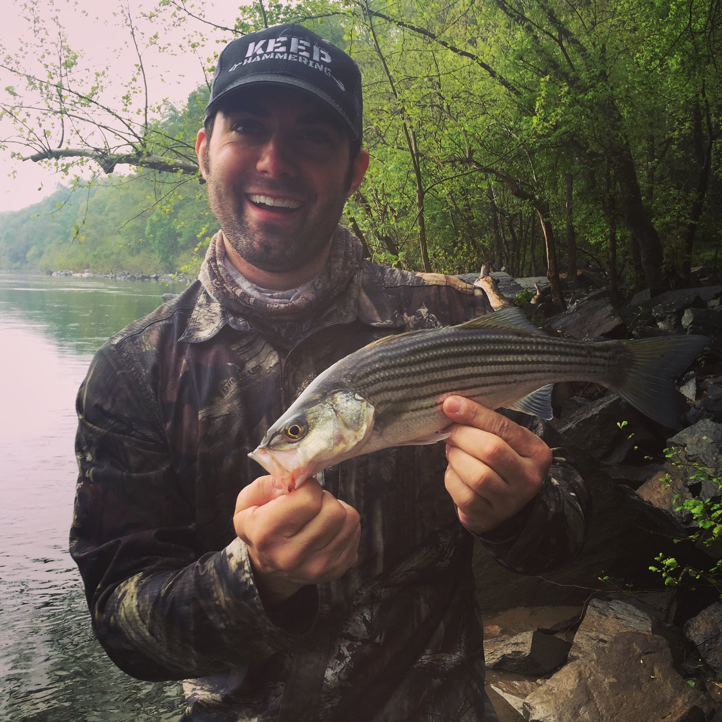 A man in camouflage clothing and a cap holding a striped bass fish by the water's edge with trees in the background.