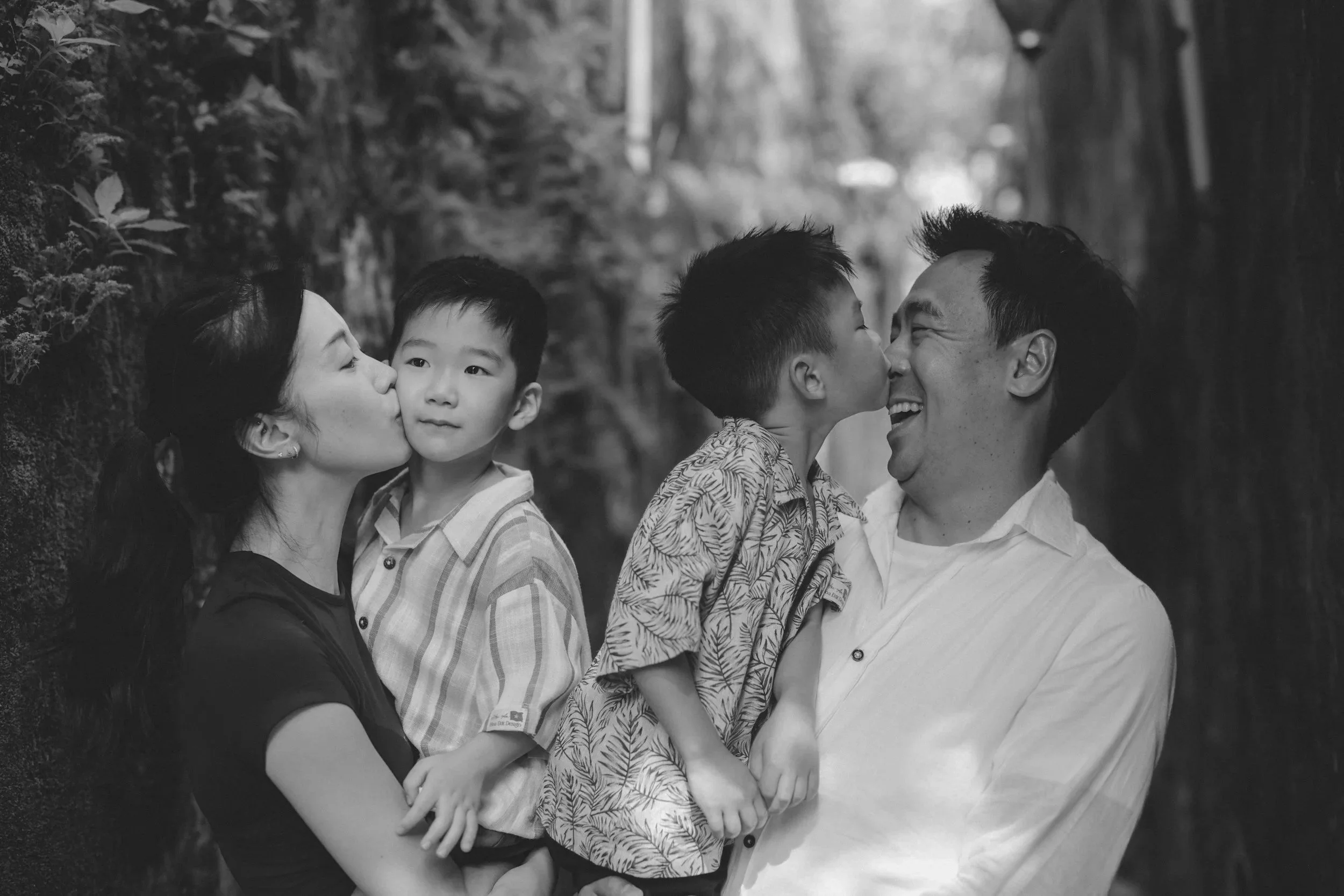 A black-and-white photo of a family, including a woman, two young boys, and a man, standing outdoors near a fence and trees. The woman and the two boys are kissing the man on the cheeks, and they are all smiling.