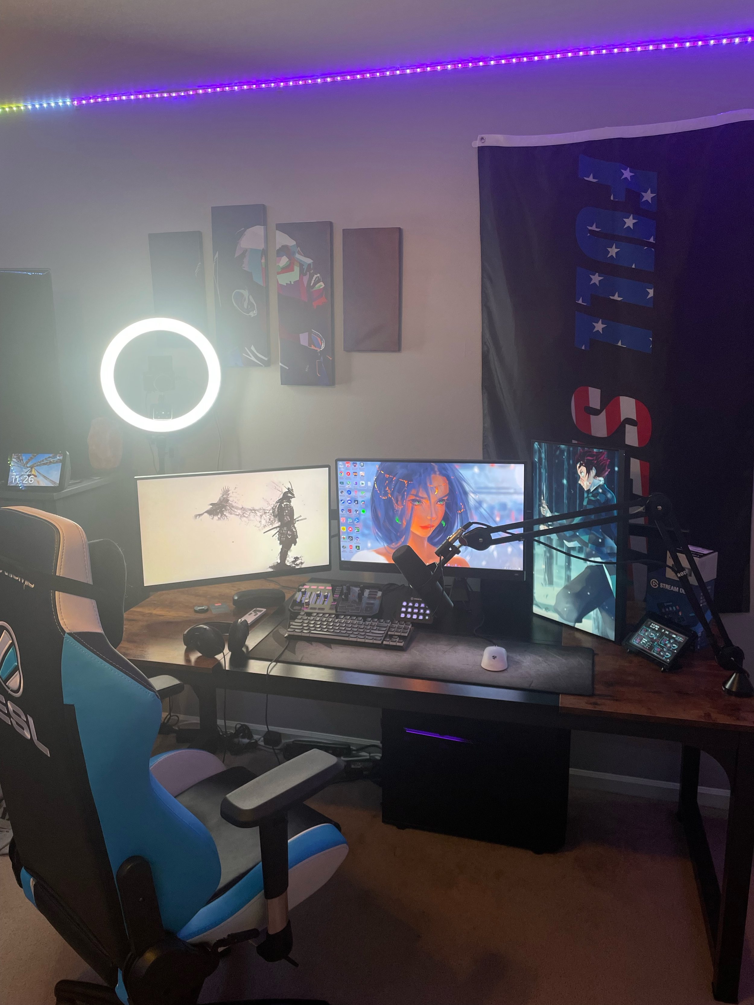 A gaming or streaming setup with three monitors, a ring light, a microphone, a keyboard, mouse, headphones, and a gaming chair in front. There are colorful LED strip lights on the ceiling and a large black flag with