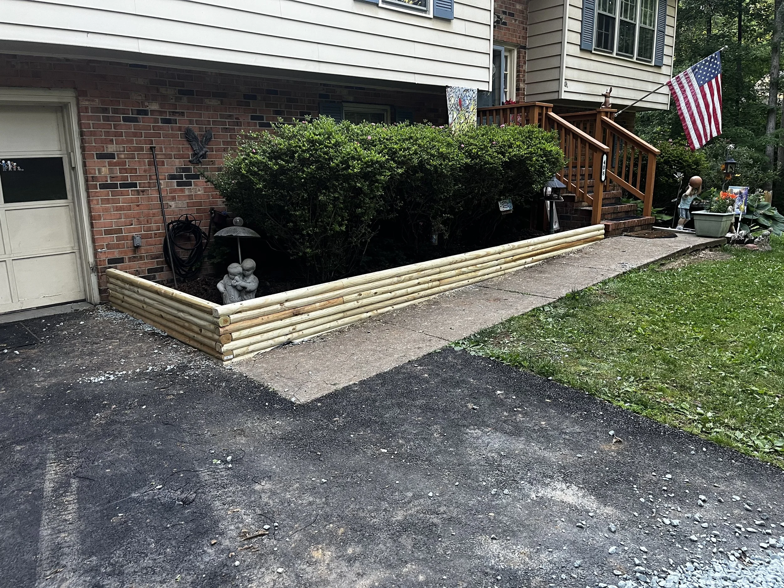 Front yard with a raised flower bed bordered by logs, featuring a garden statue, greenery, decorative items, and an American flag attached to the wooden stairs of a house with a brick and siding exterior.