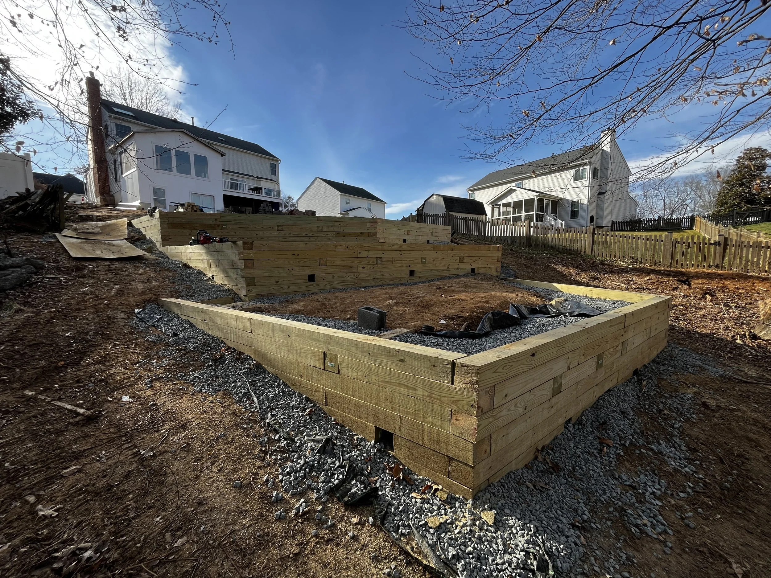 A wooden picnic table with attached benches outdoors on grass, surrounded by greenery.