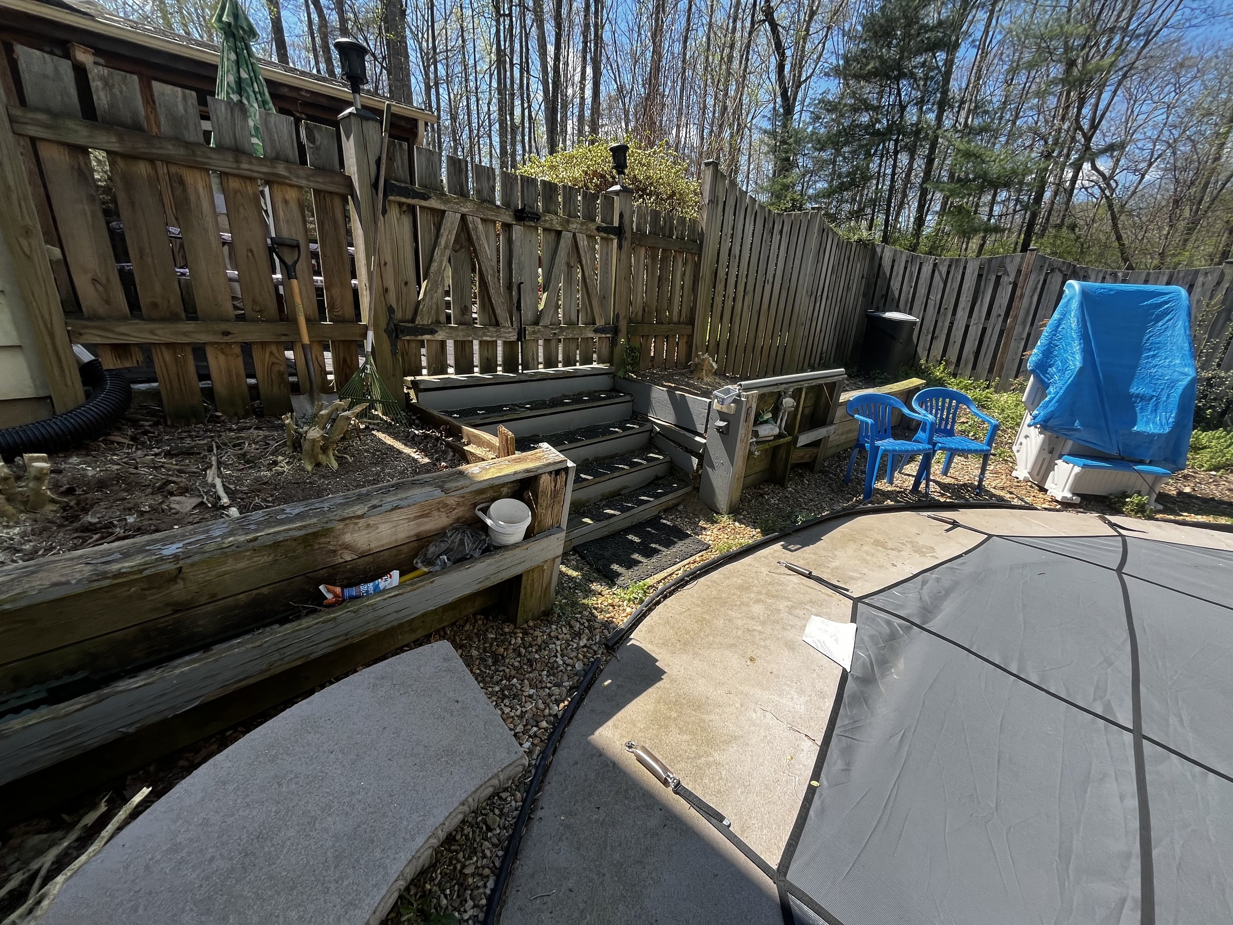 A backyard patio area with a wooden fence, some garden plants, a small set of stairs, outdoor chairs, a covered item, and a semi-circular concrete patio surface.