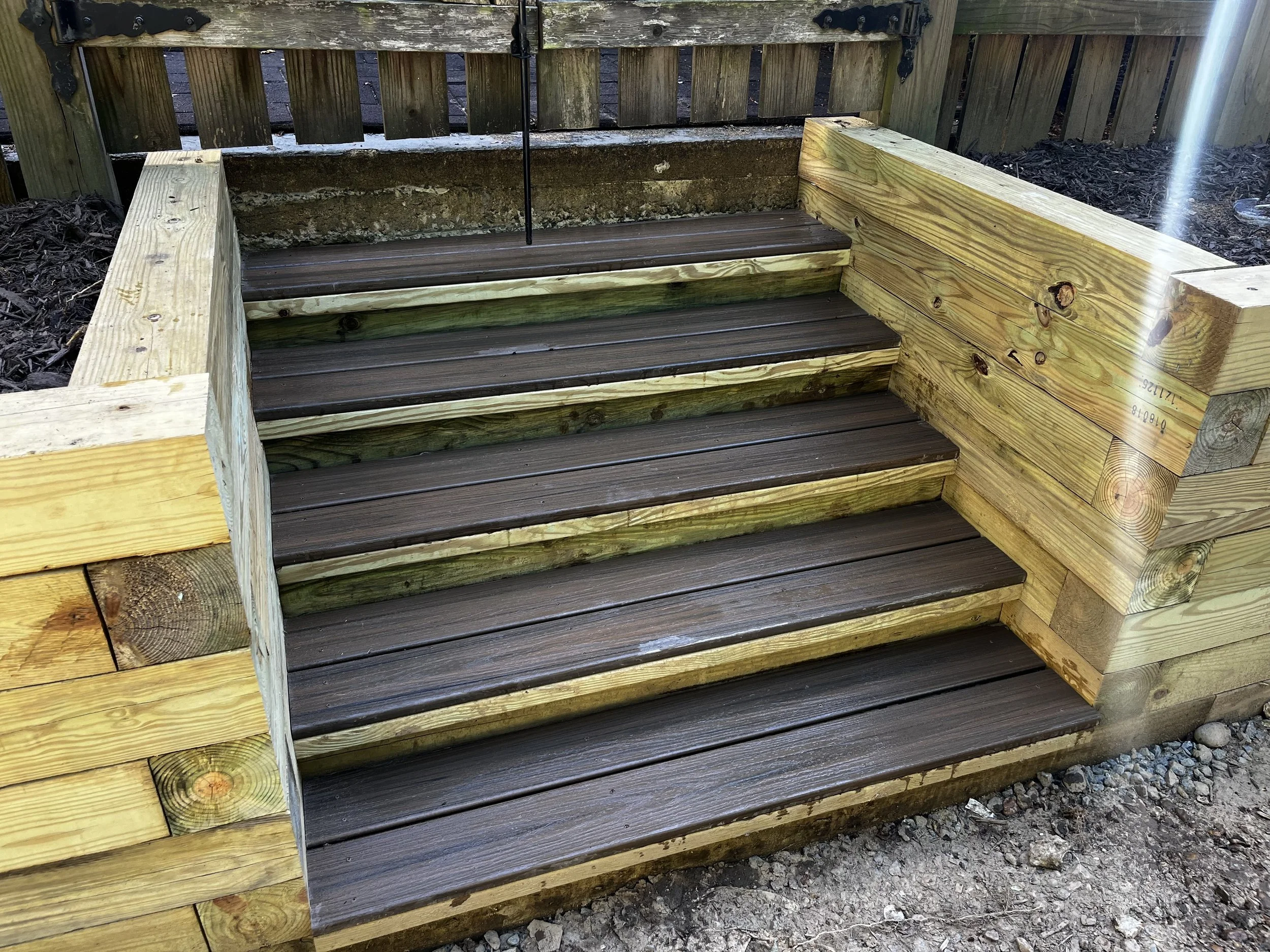 Wooden outdoor staircase with dark stained steps and light unfinished side panels, surrounded by mulch and gravel, with a wooden fence in the background.