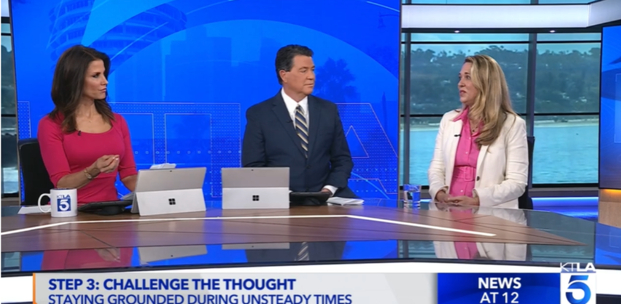 TV news segment with three anchors sitting at a curved table, two women and one man, with news graphics and a window showing water and trees in the background.
