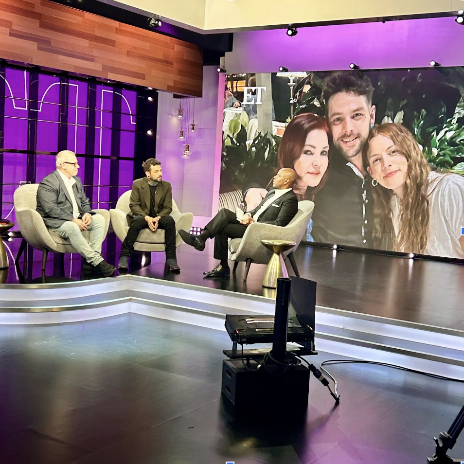 Three men are seated in chairs on a television set stage with purple and wood decor. They are engaged in a discussion, with a large screen behind them displaying a photo of three people smiling, outside the ET logo.