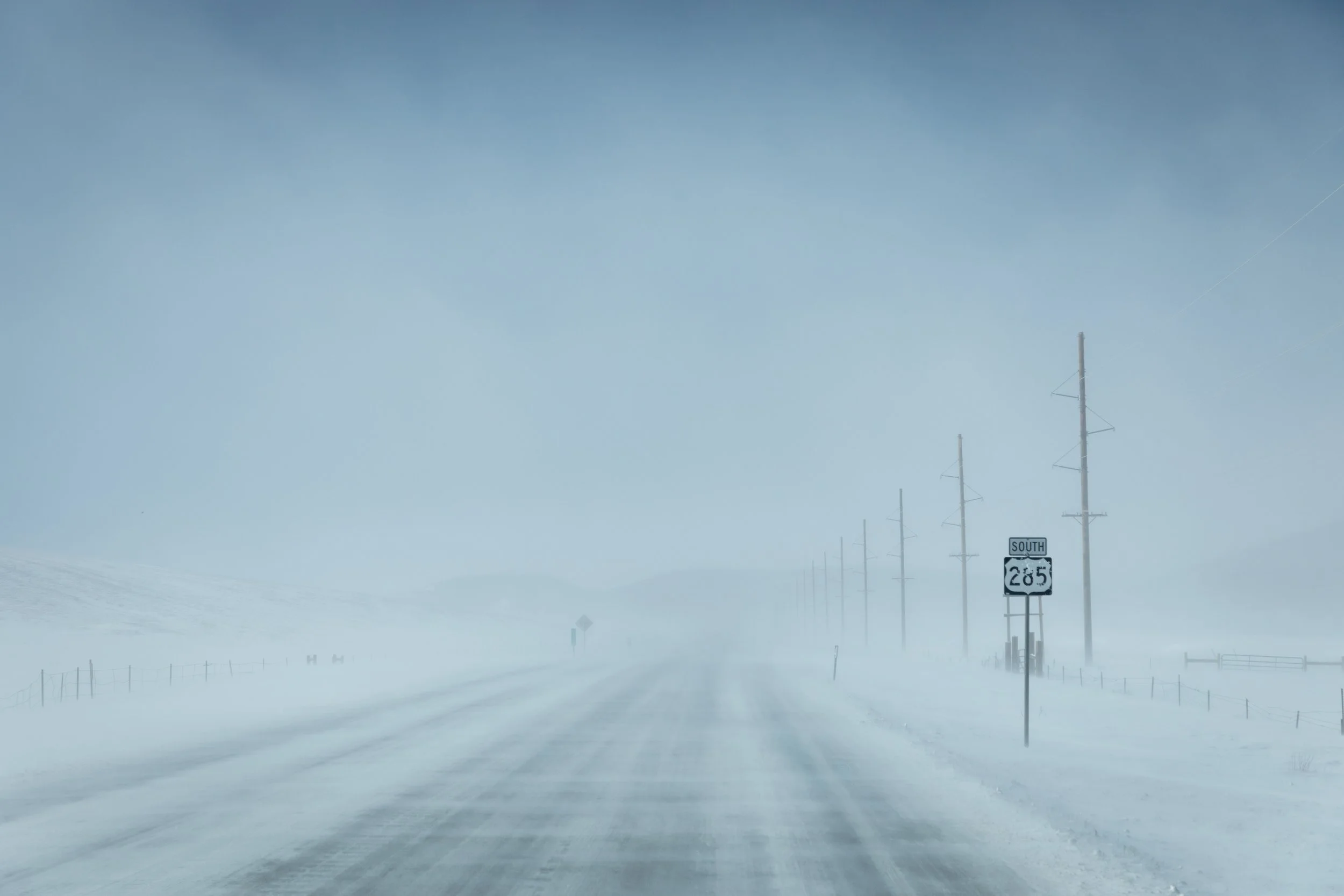 A snow-covered highway with utility poles along the side, a road sign indicating Route 285 South, and a foggy, overcast sky.