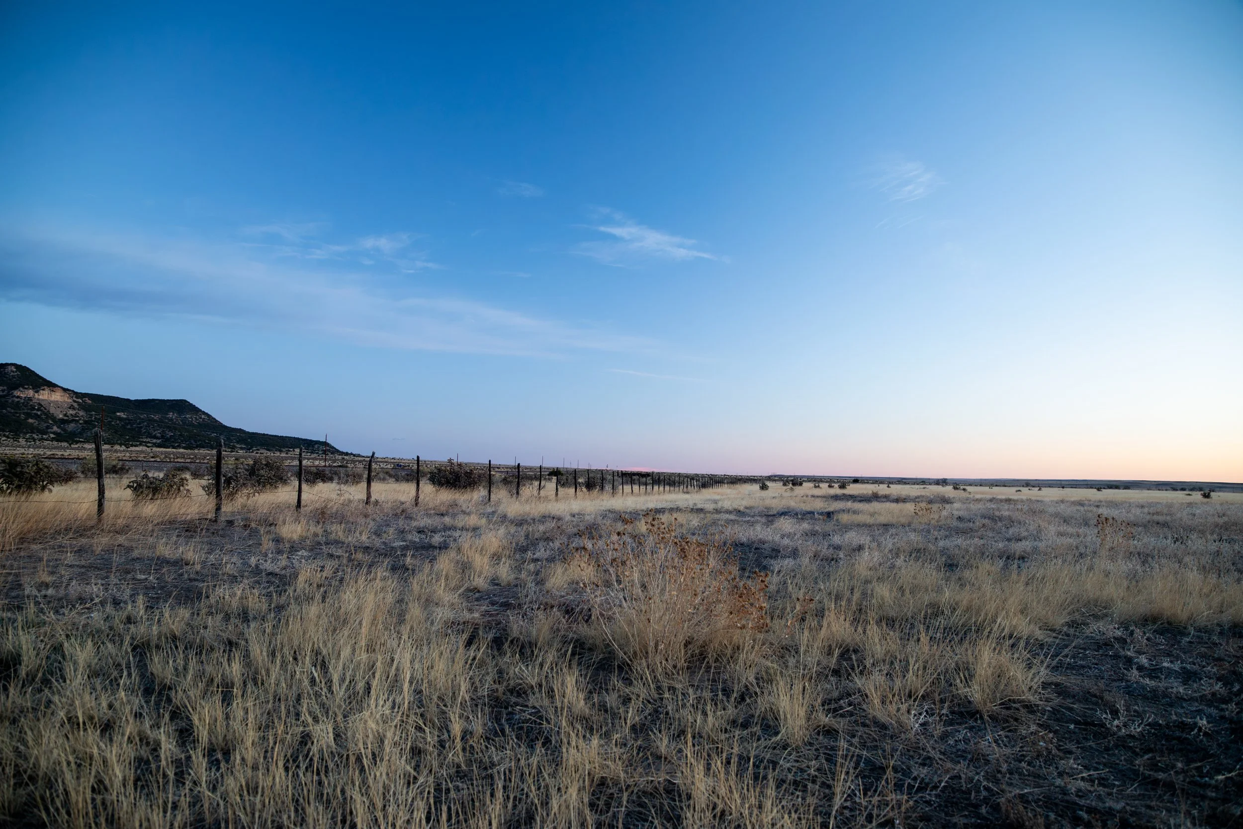 Wide view of a rural landscape with dry grass, a barbed wire fence, hills in the background, and a clear blue sky.