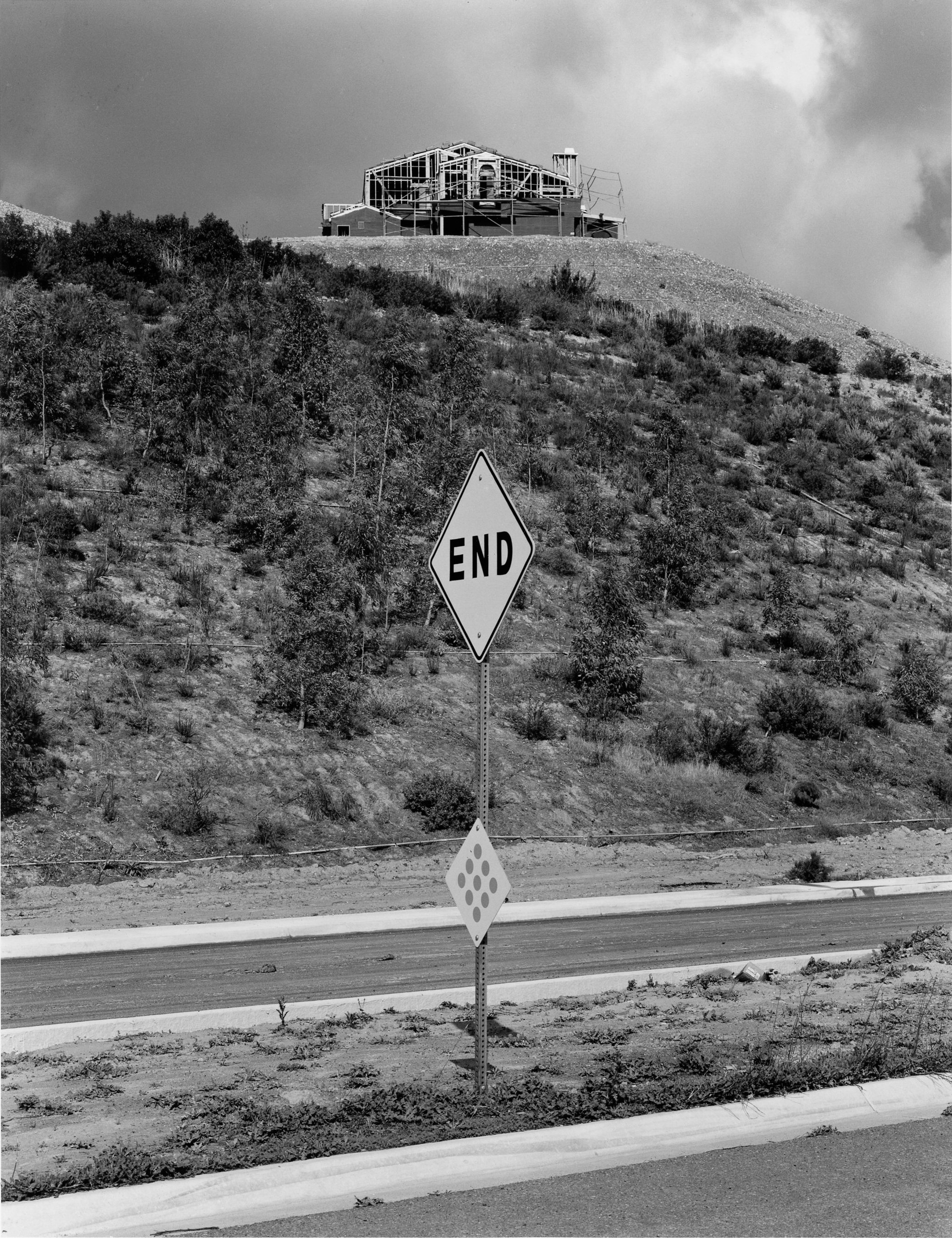 A black and white photo of a house on a hill under construction, with scaffolding visible around it. In the foreground, there is a sign that says 'END' and a smaller sign with circular patterns, along a roadside.