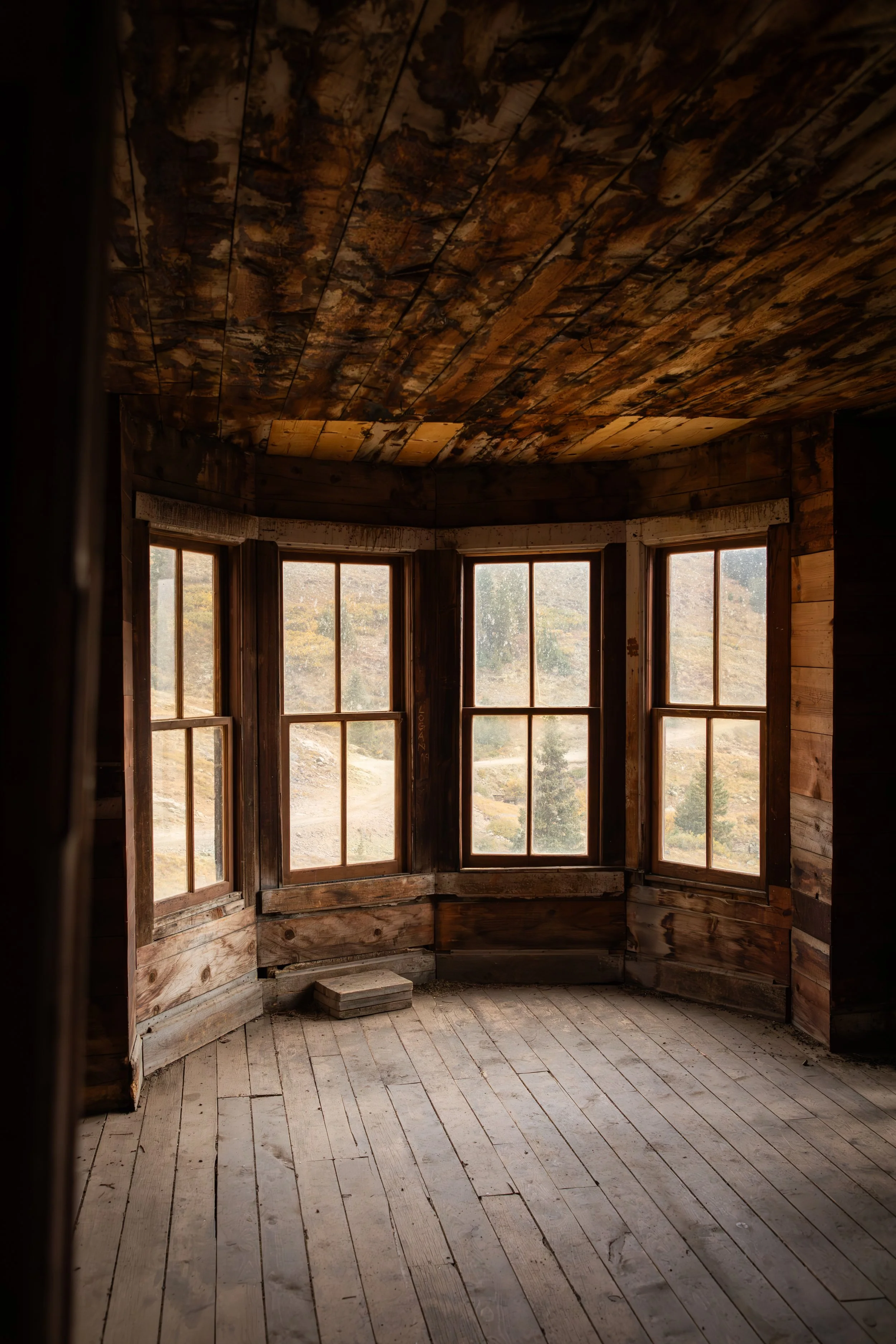 Interior of a rustic wooden cabin with large bay windows looking out onto a landscape of trees and hills, unfinished wooden floor, and a weathered wood ceiling.
