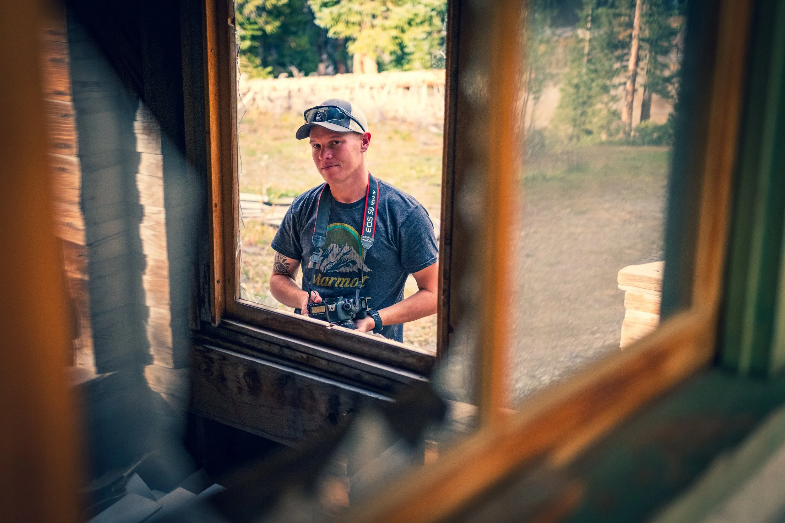 A man wearing a baseball cap, sunglasses on top, and a graphic t-shirt holding a camera, seen through a small rectangular window, outdoors with trees in the background.
