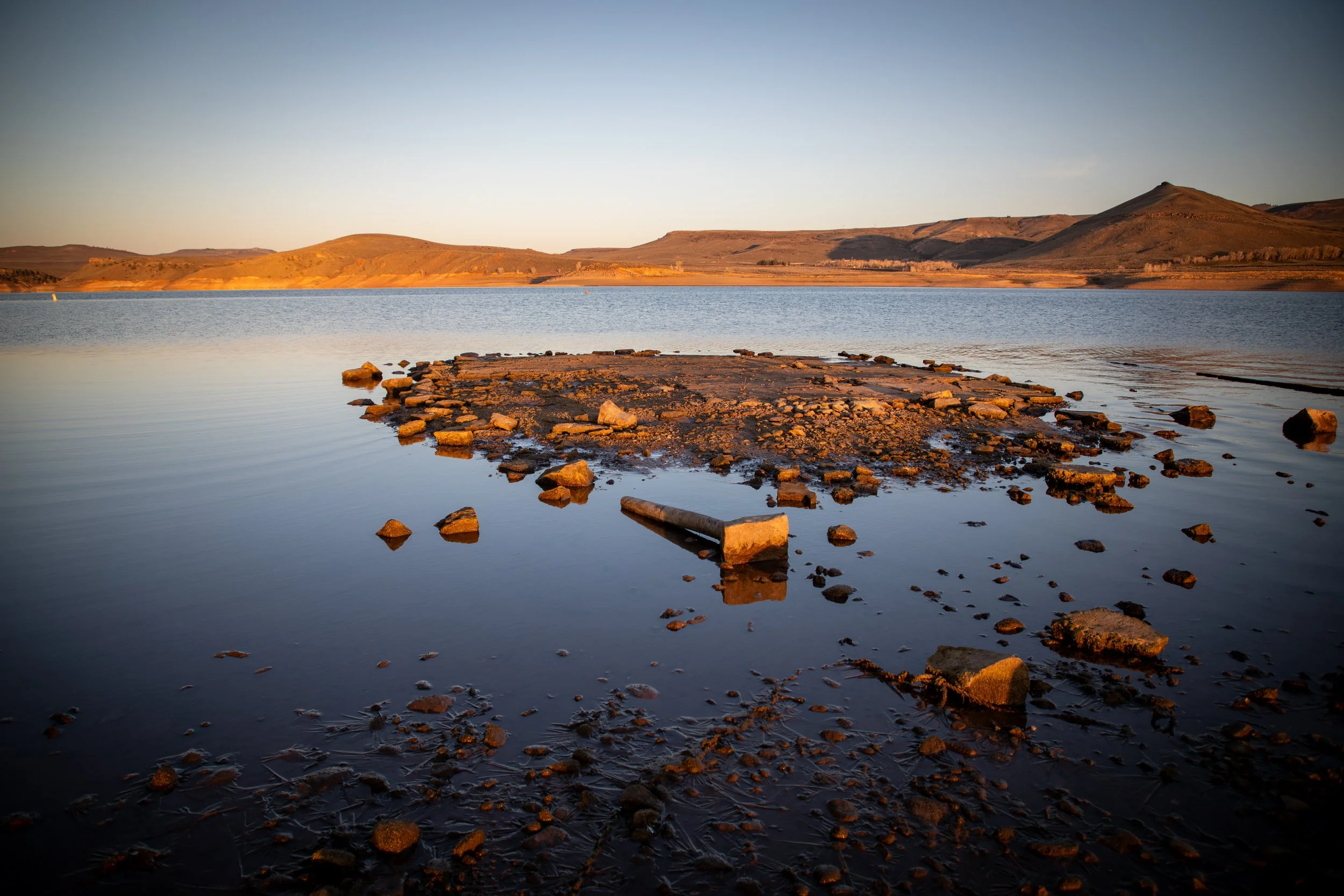 A lake with a rocky shoreline and distant hills under a clear sky at sunset.