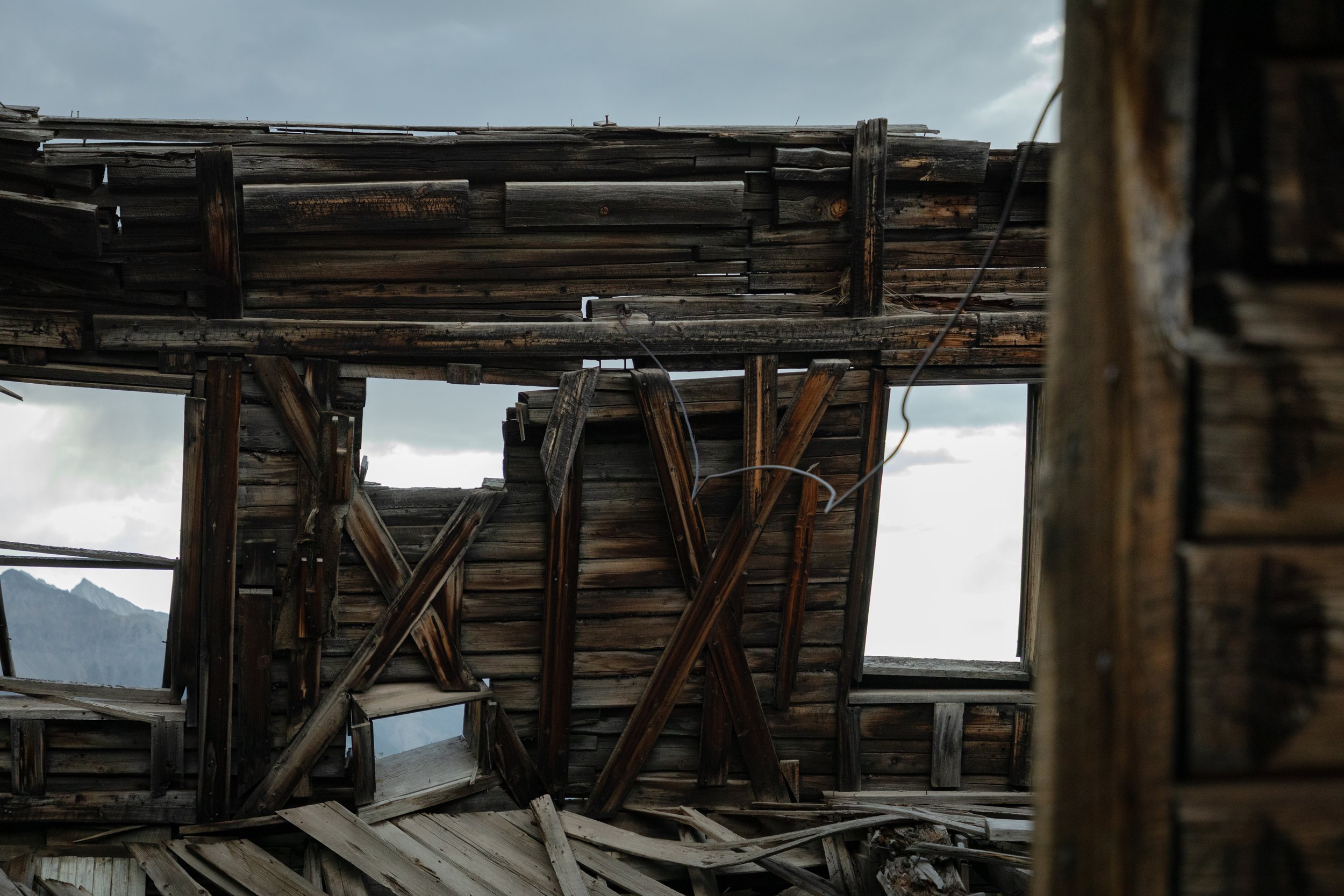 The image shows the remains of a collapsed wooden structure, with broken and charred wood, against a cloudy sky and mountains in the background.