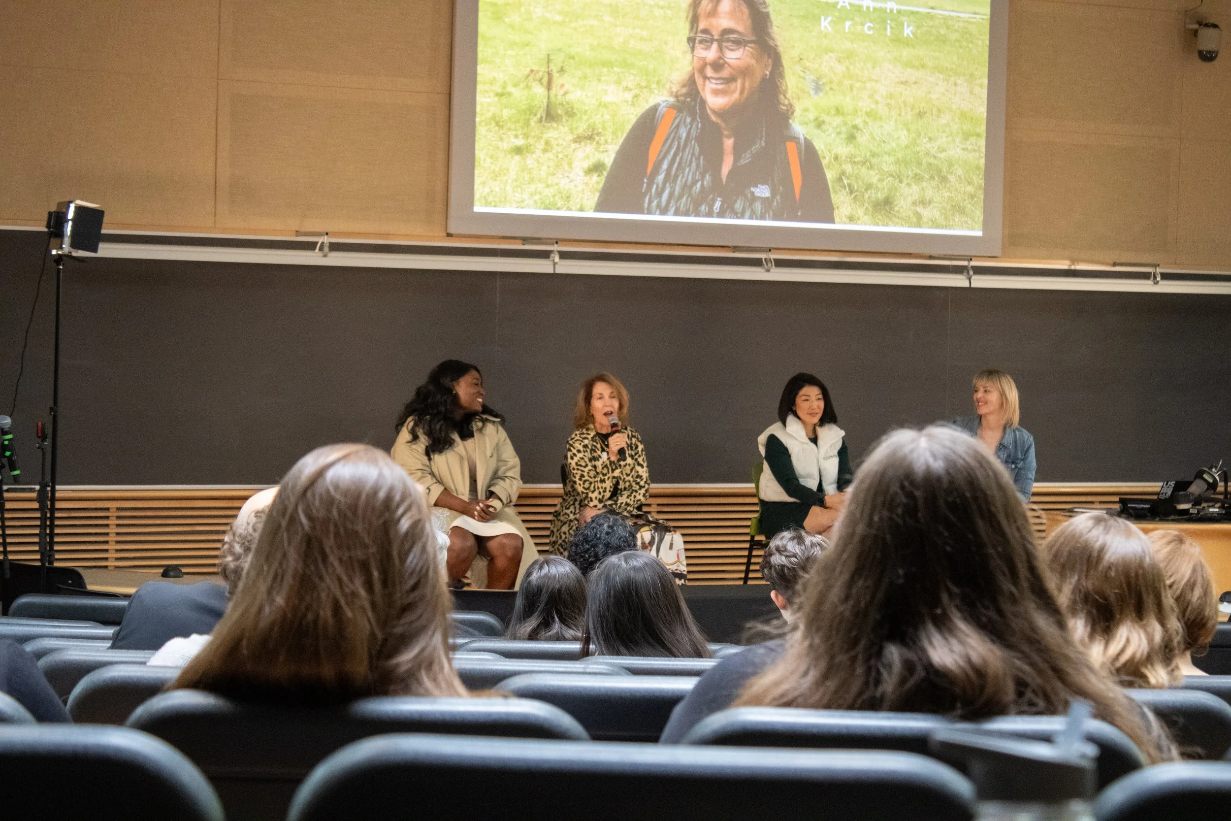 Panel discussion with four women in an auditorium, audience seated, screen above shows a woman outdoors with glasses and a scarf.