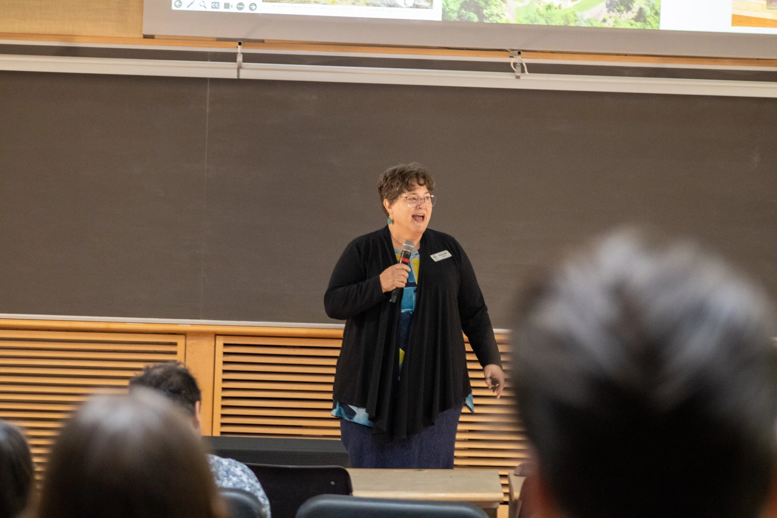 A woman with short curly hair, glasses, and a name tag is speaking into a microphone in a classroom or lecture hall, with students seated in front of her. She appears engaged and is standing in front of a blackboard.