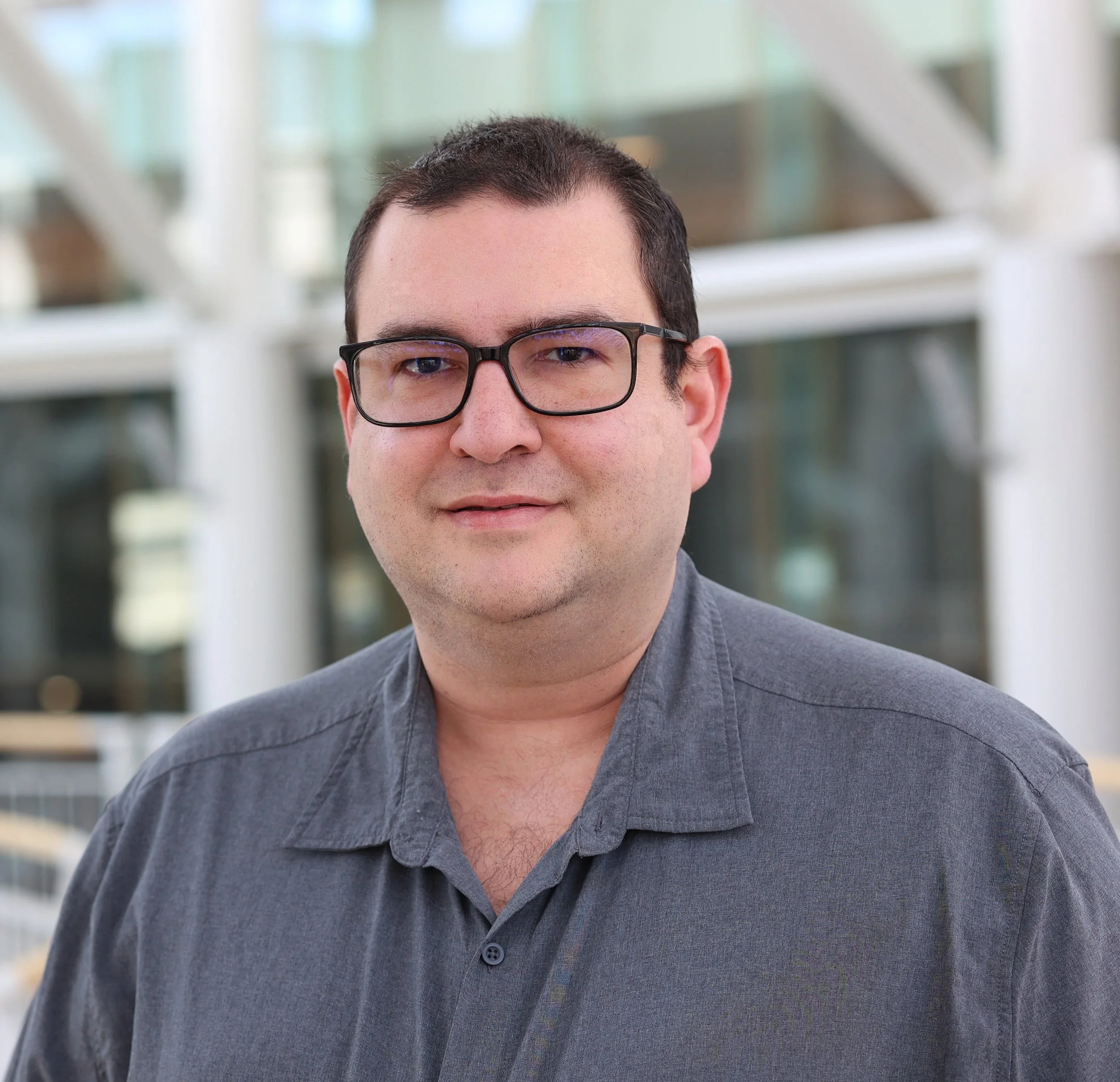 A man with short dark hair, glasses, and a gray shirt standing outdoors near a modern glass building.