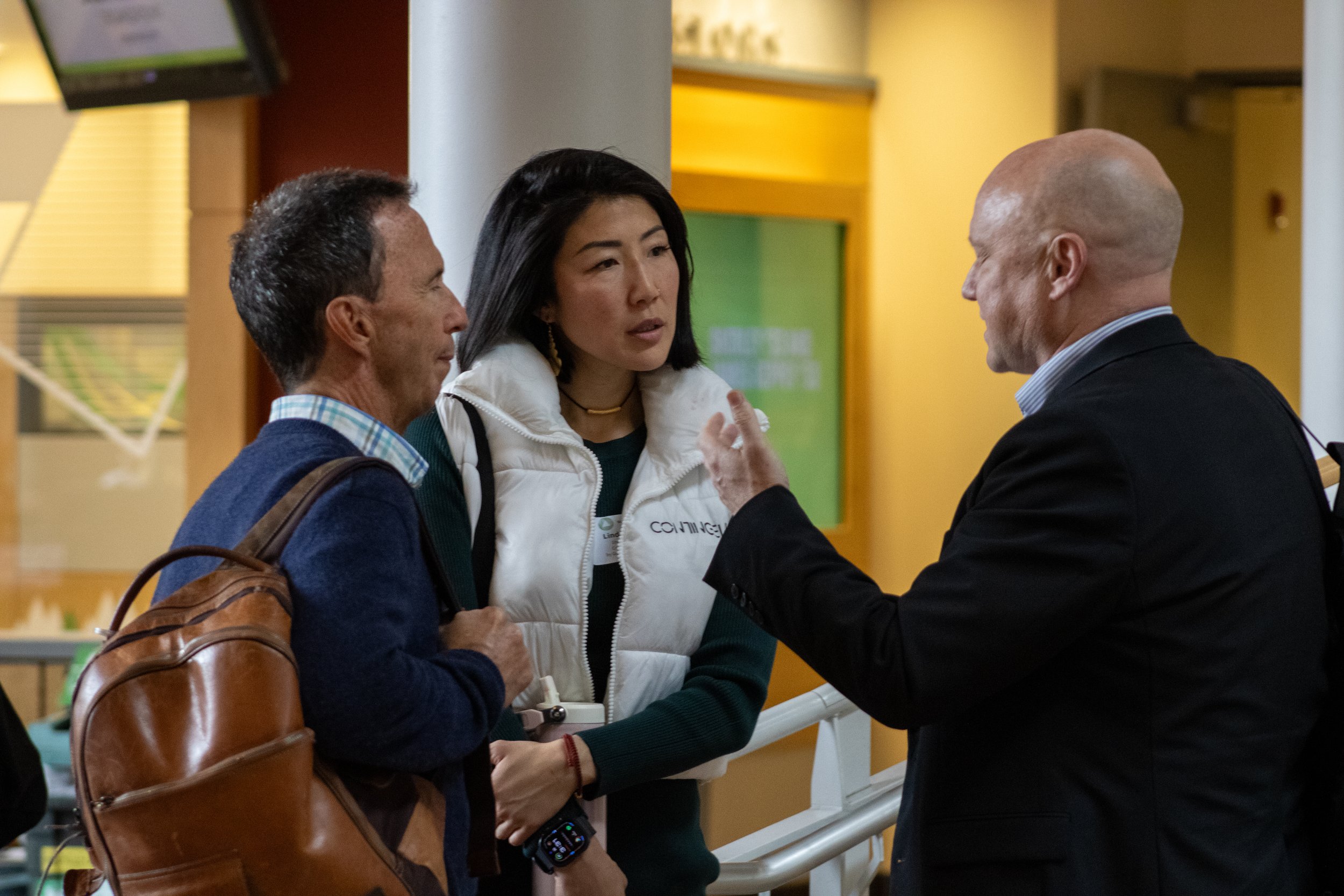 Three people engaged in conversation at an indoor location, including a man with a backpack, a woman wearing a white vest, and a bald man in a suit.
