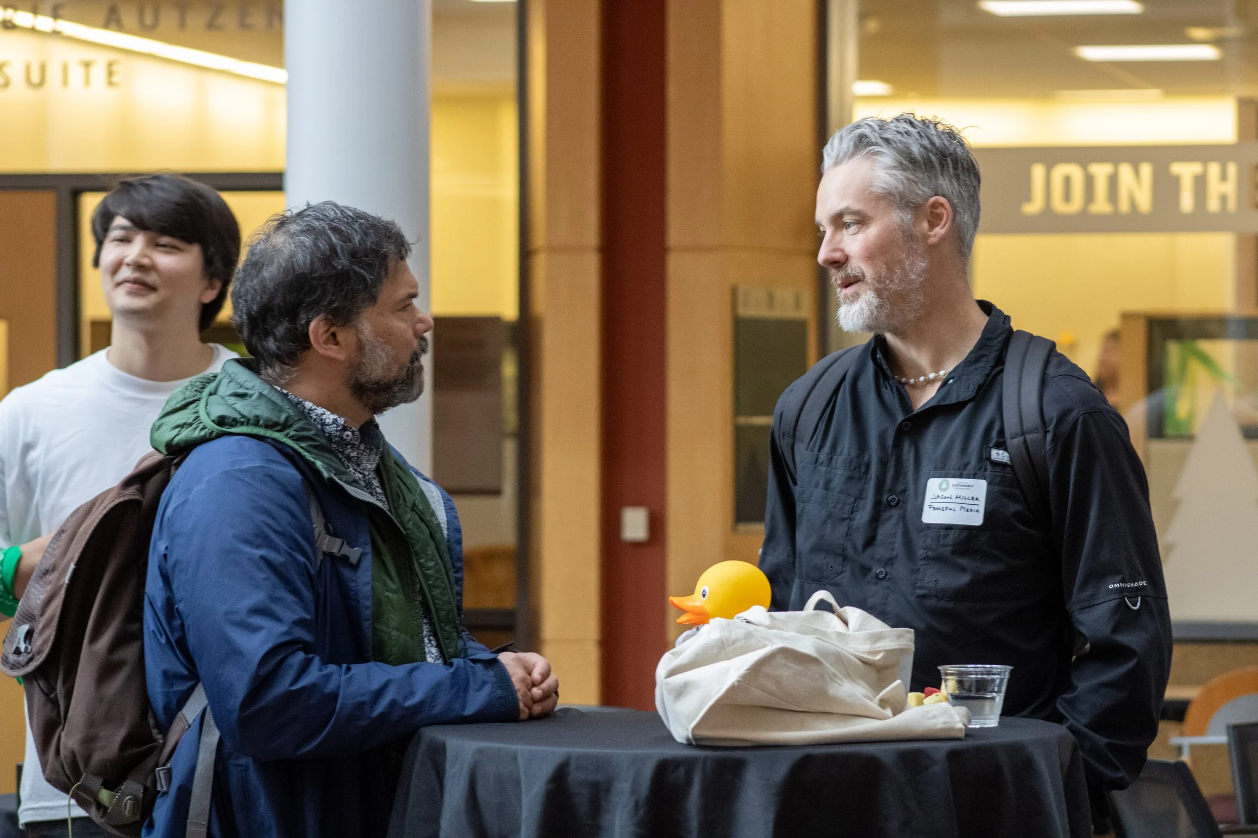Two men having conversation at a table in an indoor setting, with a rubber duck and a bag on the table, and a young man standing in the background smiling.