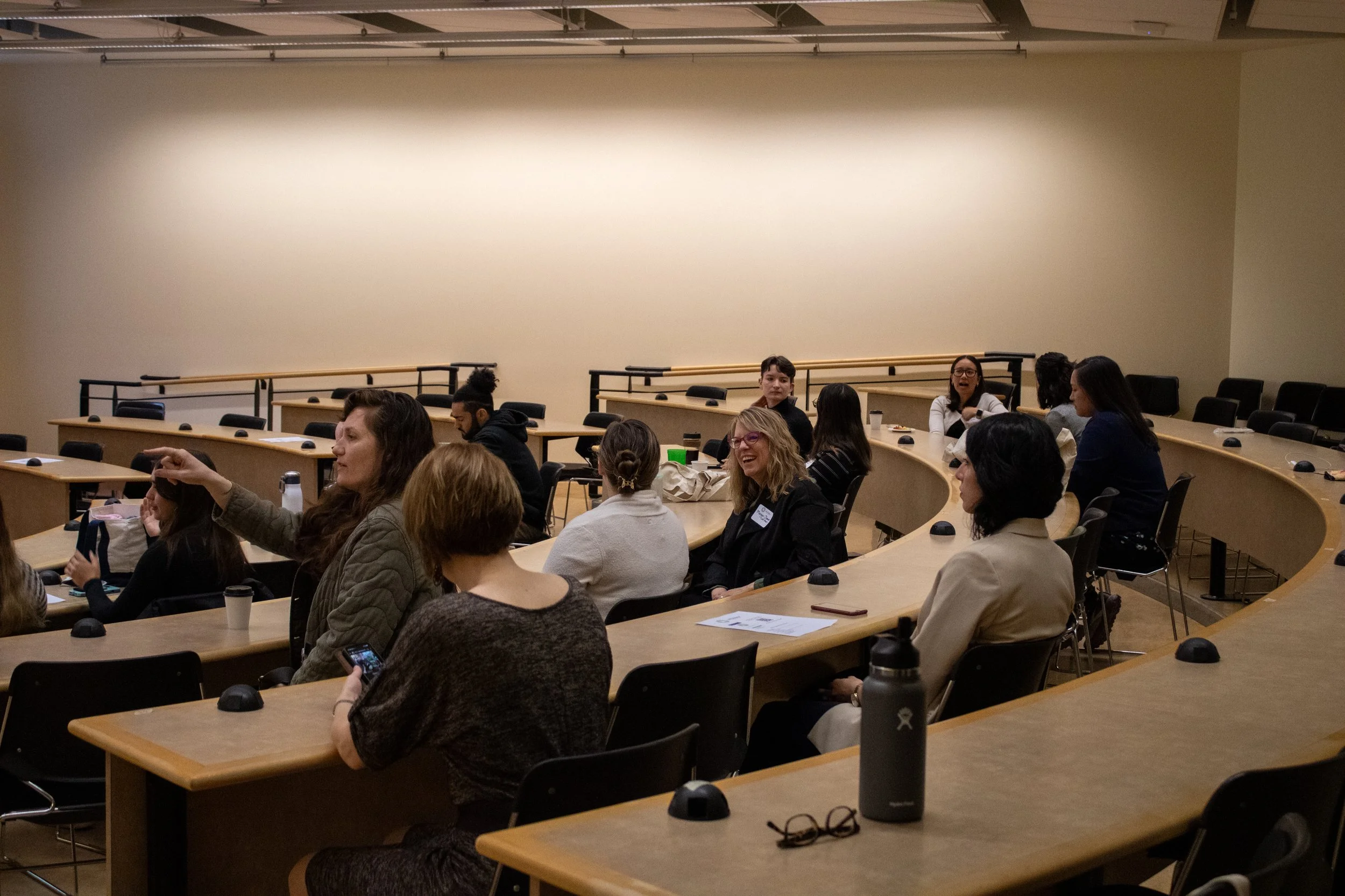 People attending a lecture or conference in a tiered classroom setting, some engaged in conversation, others focused on a speaker or presentation.