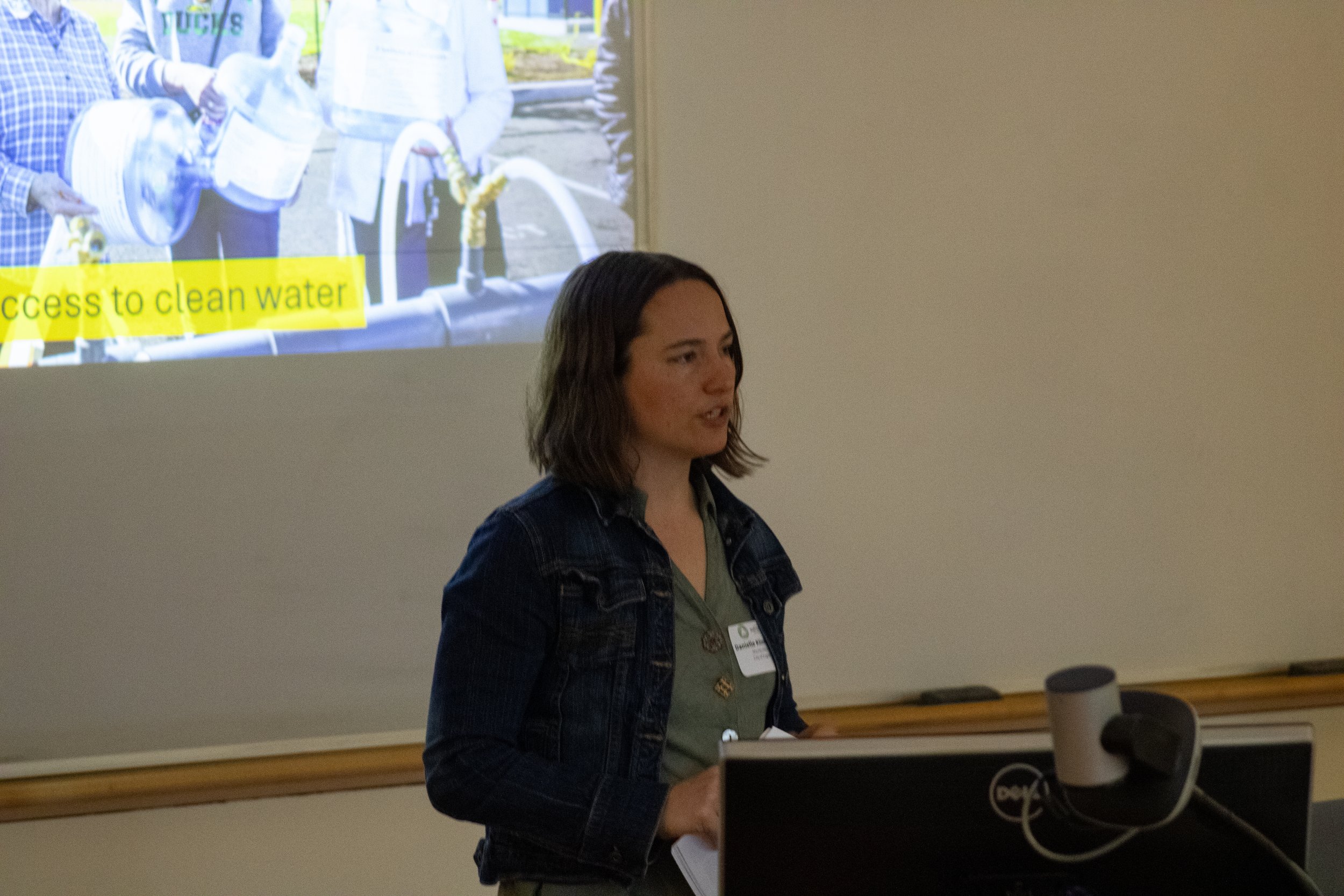 A woman giving a presentation in a classroom or conference room, with a slide projected behind her showing people working outdoors on a water project and the text 'access to clean water'.