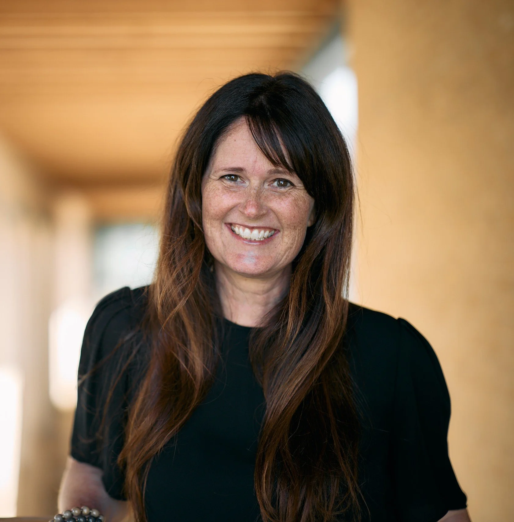 Smiling woman with long brown hair and freckles, wearing a black top, standing in a blurred indoor hallway.