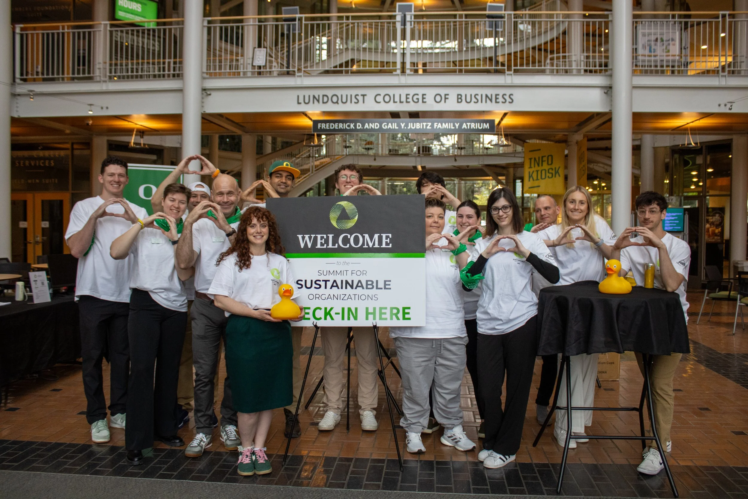 Group of people standing behind a sign that says 'Welcome to the Summit for Sustainable Organizations, Check-in Here' at Lundquist College of Business. Some are making heart shapes with their hands, and two of them are holding yellow rubber duckies.