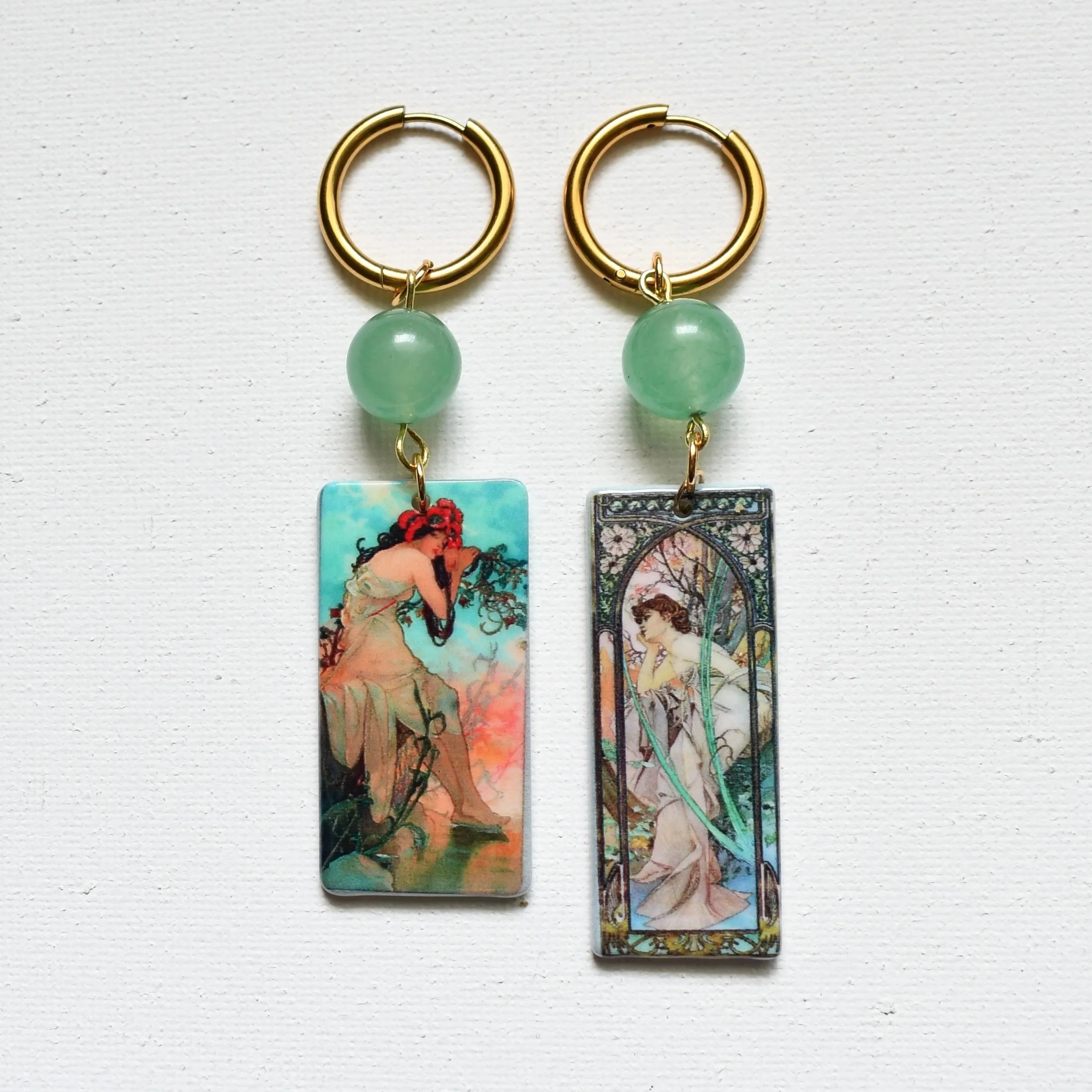 Pair of gold hoop earrings with large green beads and rectangular pendants, each featuring a different artistic illustration of women.