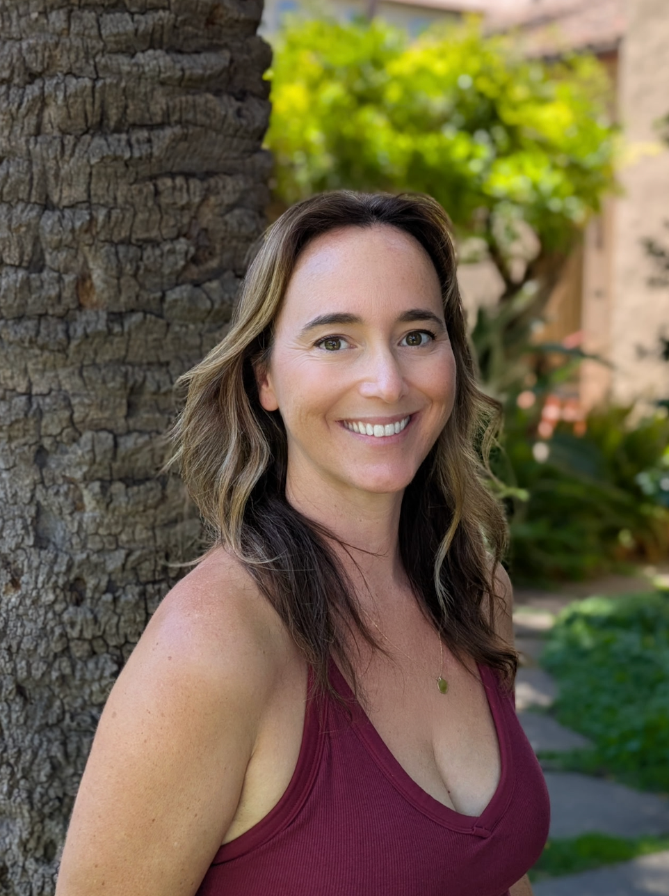 A woman with shoulder-length wavy brown hair smiling outdoors, wearing a sleeveless maroon top and a delicate gold necklace, standing near a tree with a textured bark trunk and green foliage background.