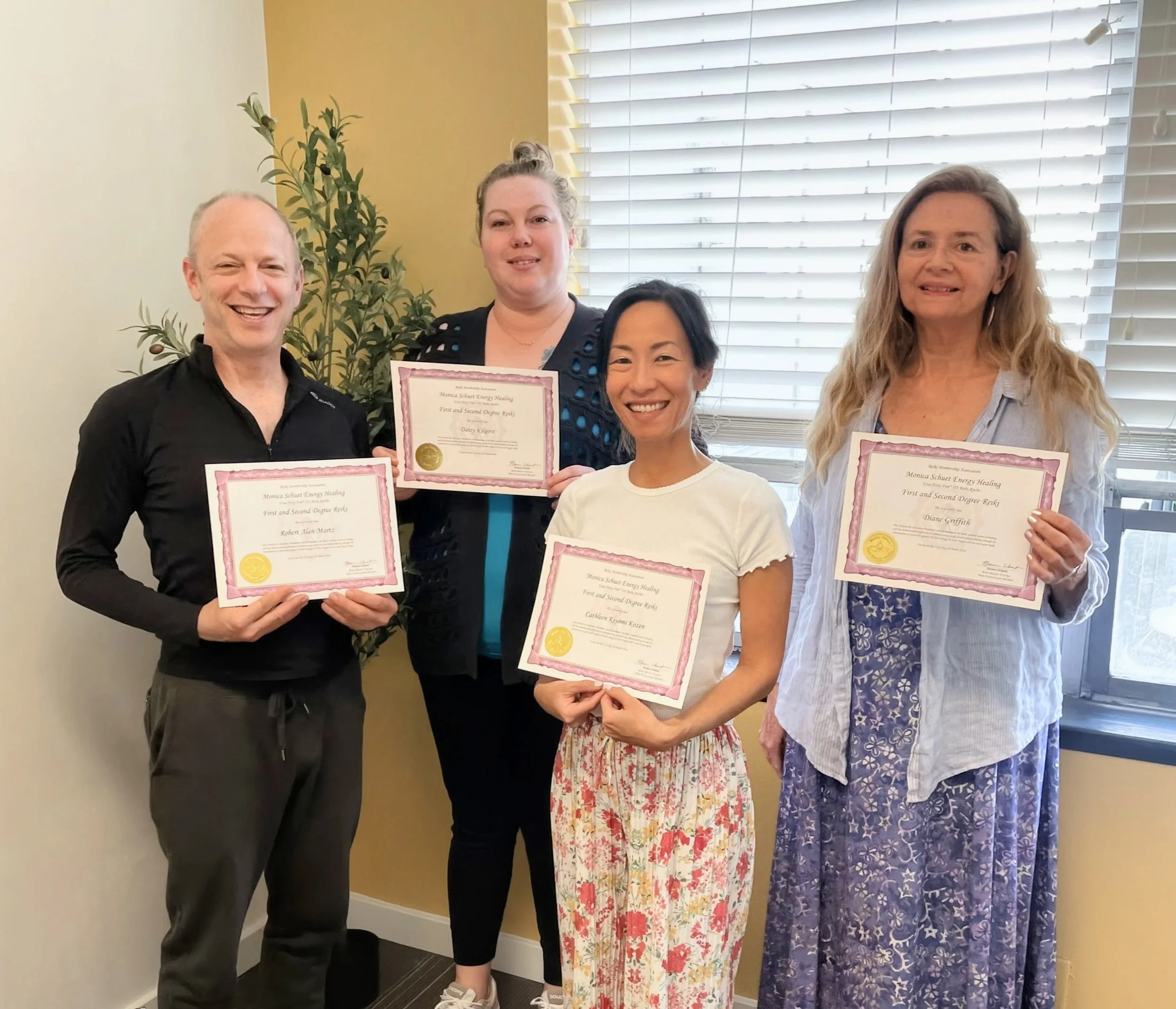 A group of four people, three women and one man, smiling and holding certificates, standing indoors near a window with blinds. One woman is wearing a floral skirt and another a pink skirt, while the man is wearing a black shirt.