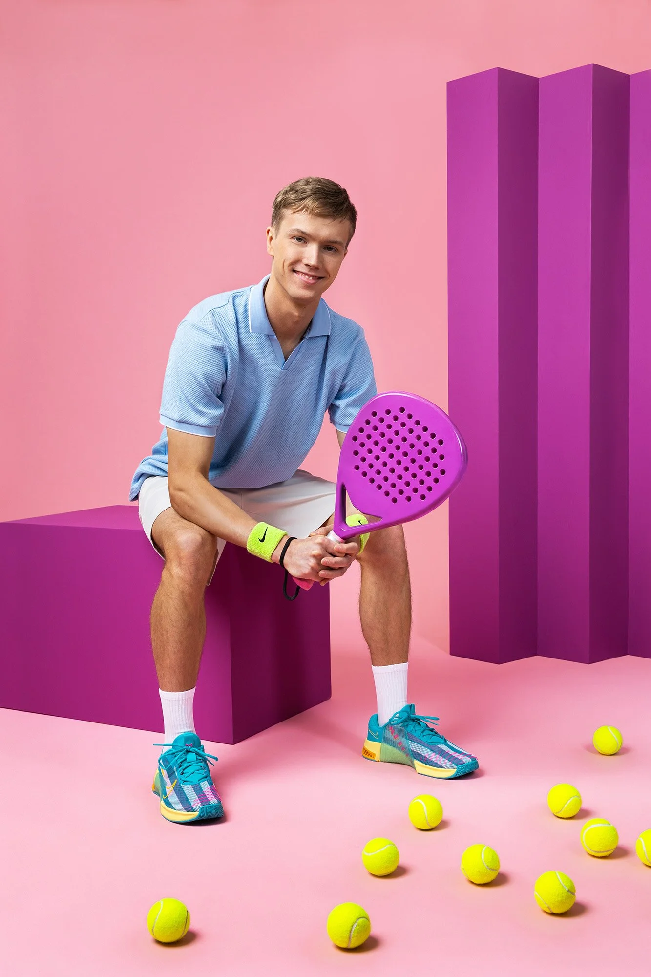 A young man dressed in tennis attire sitting on a pink block, holding a purple tennis racket, surrounded by yellow tennis balls, with a pink background and purple geometric shapes.