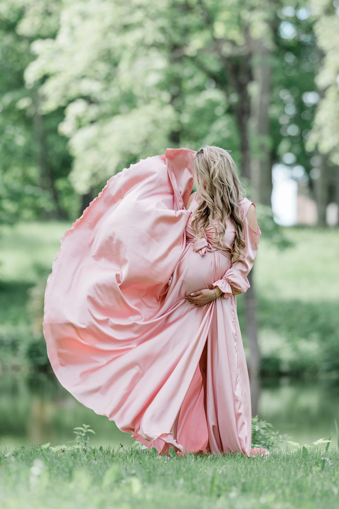 Pregnant woman in a flowing pink dress standing outdoors on a grassy area near a pond, with lush green trees in the background.