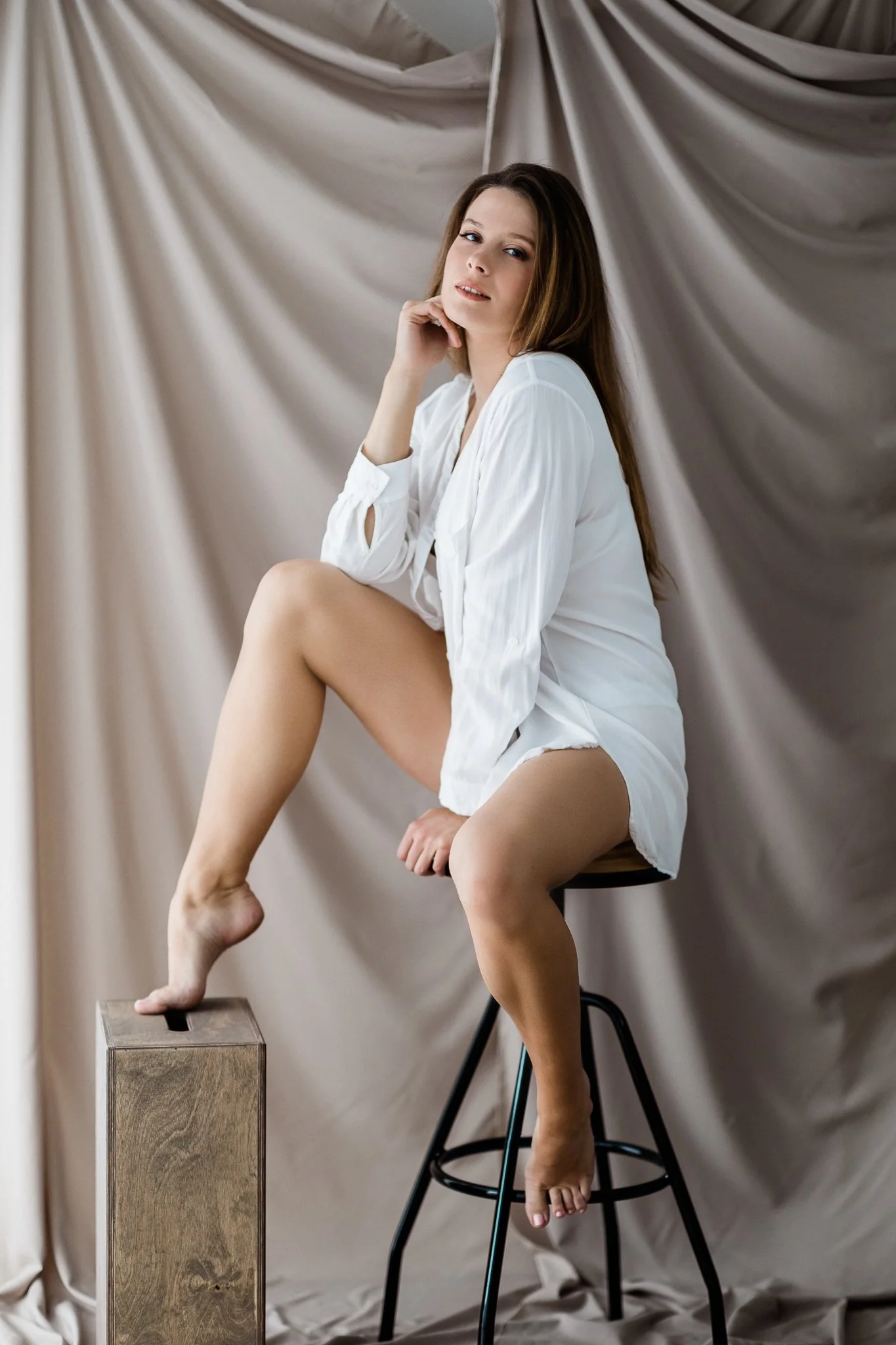 A woman with long brown hair and fair skin, sitting on a tall stool with one foot on a wooden box, wearing a white shirt and no pants, against a beige fabric backdrop.