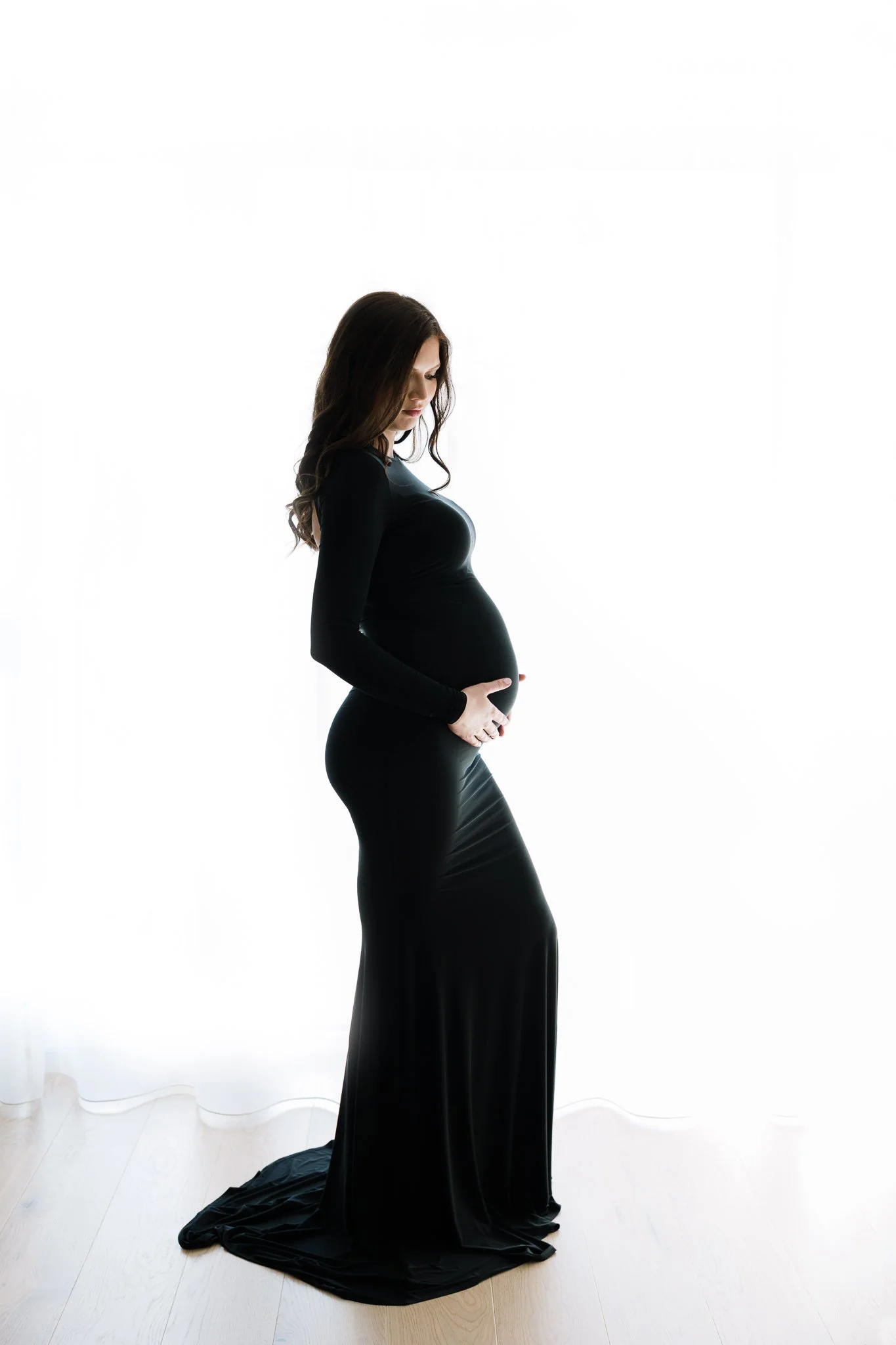 A pregnant woman in a black dress standing in front of a white background, with her hands resting on her belly and looking down.