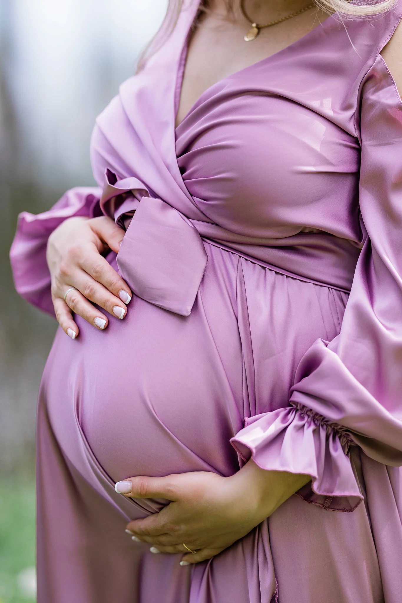 Close-up of pregnant woman's belly in a satin lavender dress, hands gently cradling her abdomen outside.