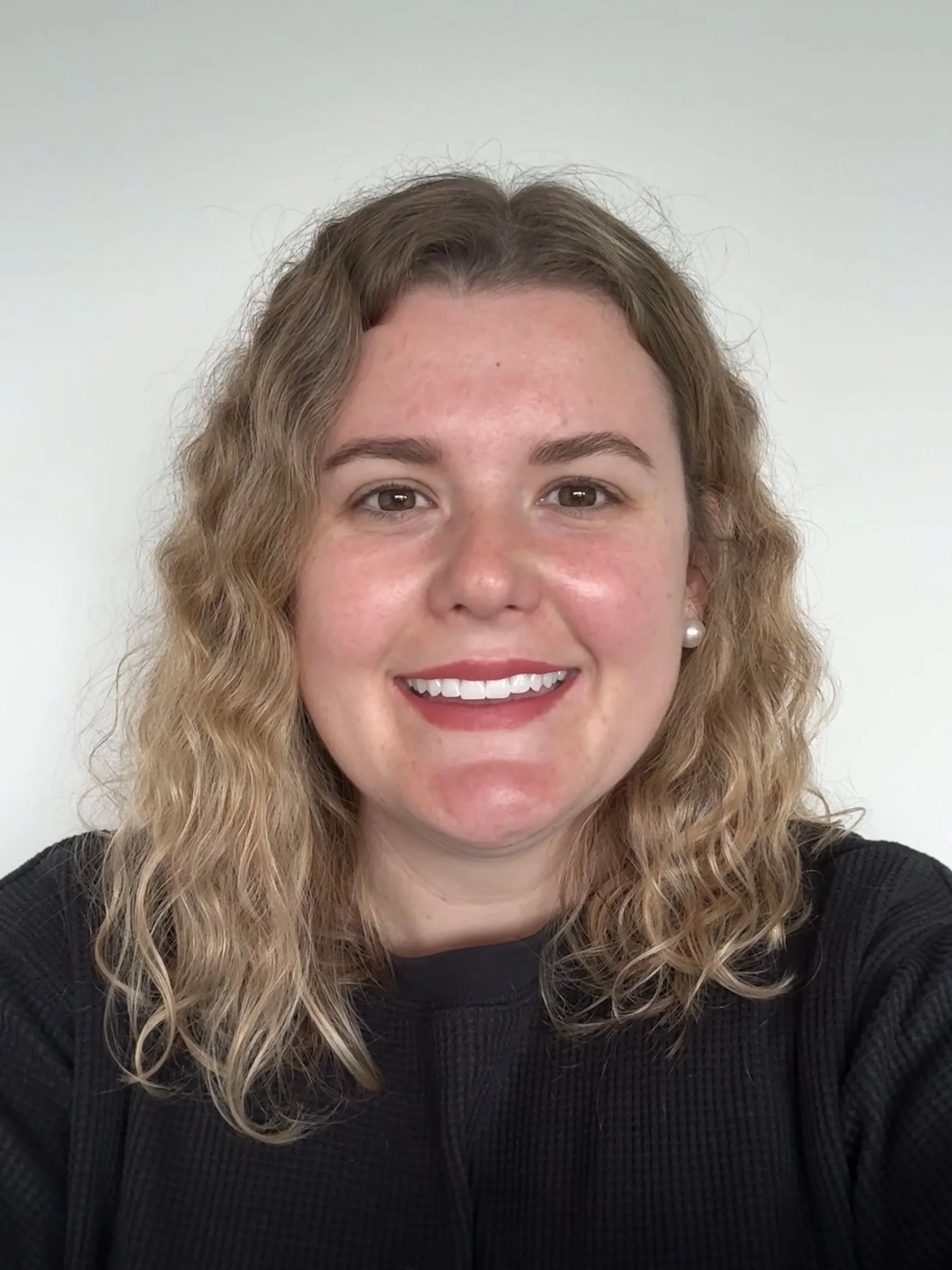 Close-up of a smiling woman with curly blonde hair, wearing a black top and pearl earrings, against a plain white background.