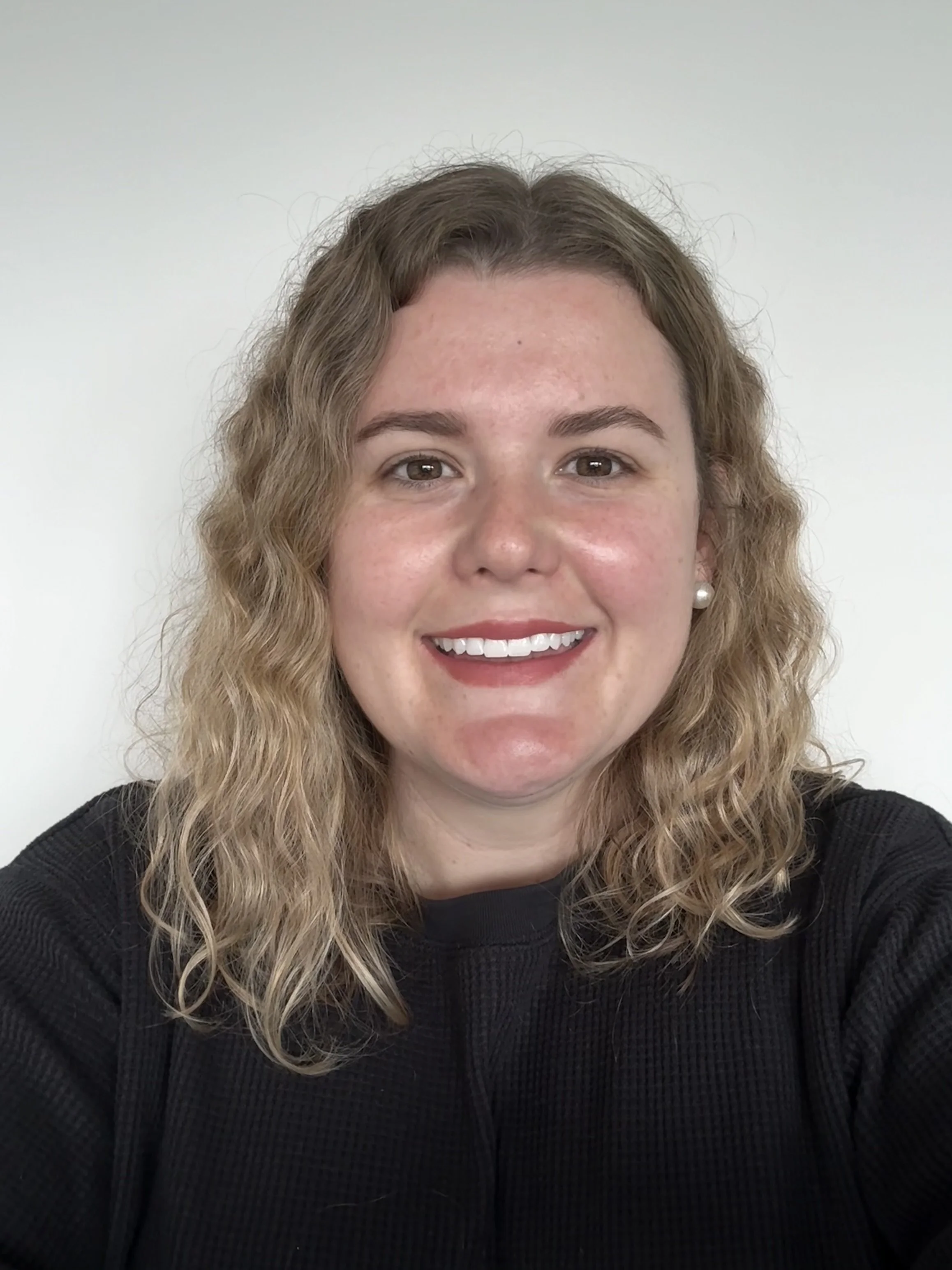 Close-up of a woman with wavy blonde hair, smiling showing her teeth, wearing pearl earrings and a black top, against a plain light-colored background.