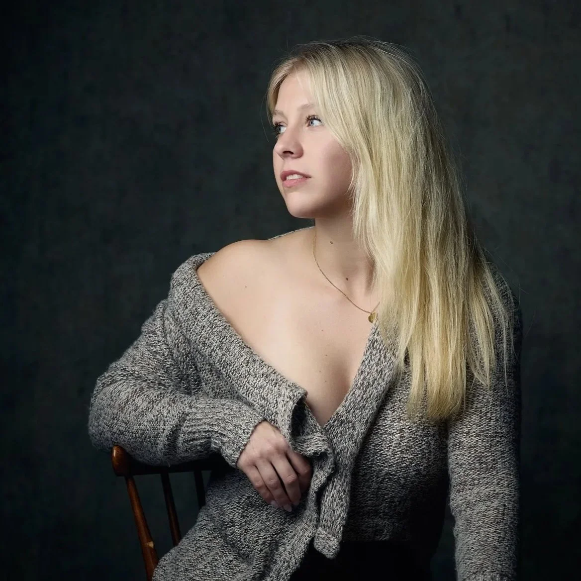 En ung kvinde med langt blond hår iført en løs grå sweater, der sidder på en stol mod en mørk baggrund.