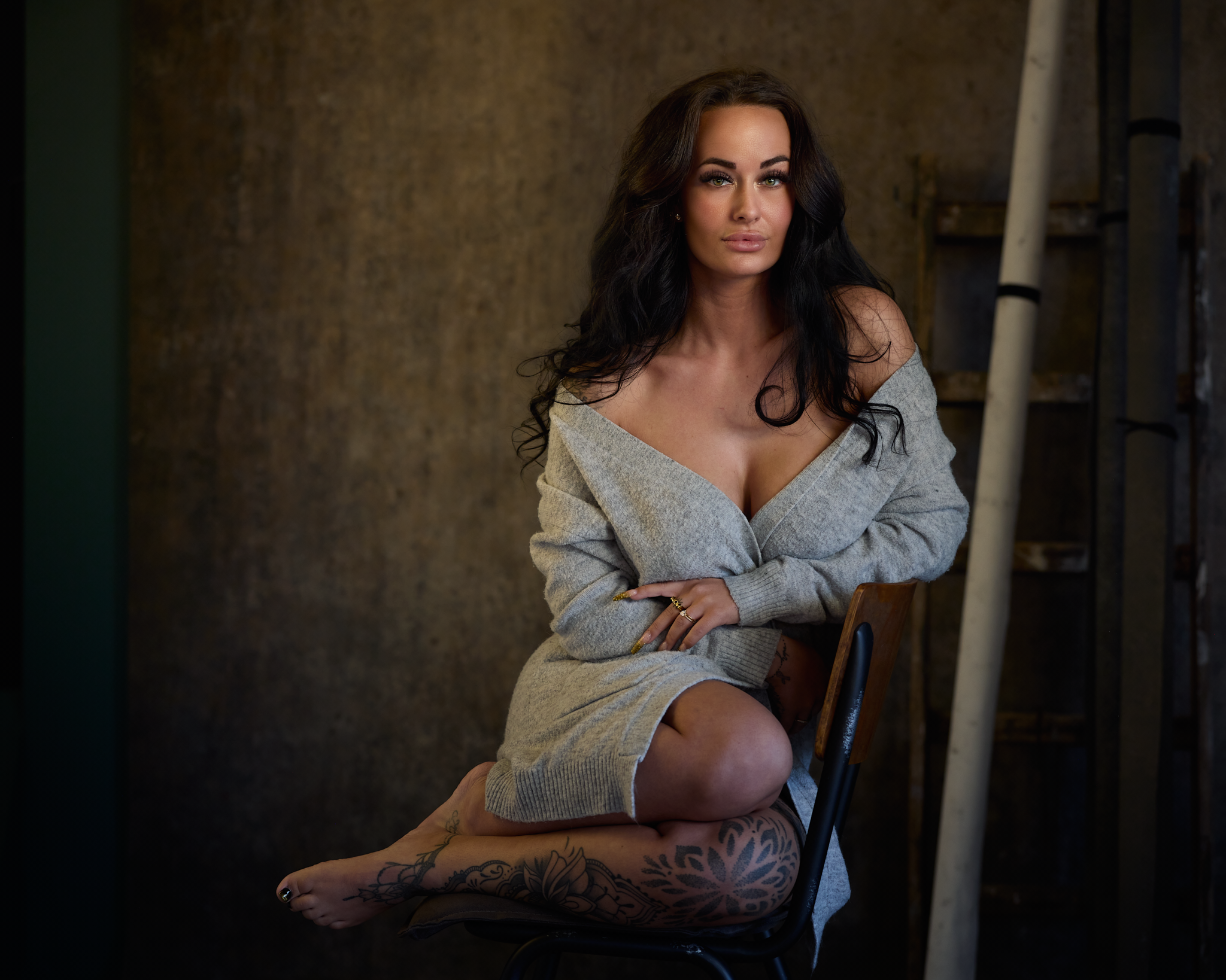 Kvindelig person med mørkt, bølget hår, iført en grå sweater klædt af skuldrene, sidder på en stol i en industriel indendørs setting med en rustik baggrund og tatoveringer på benet.