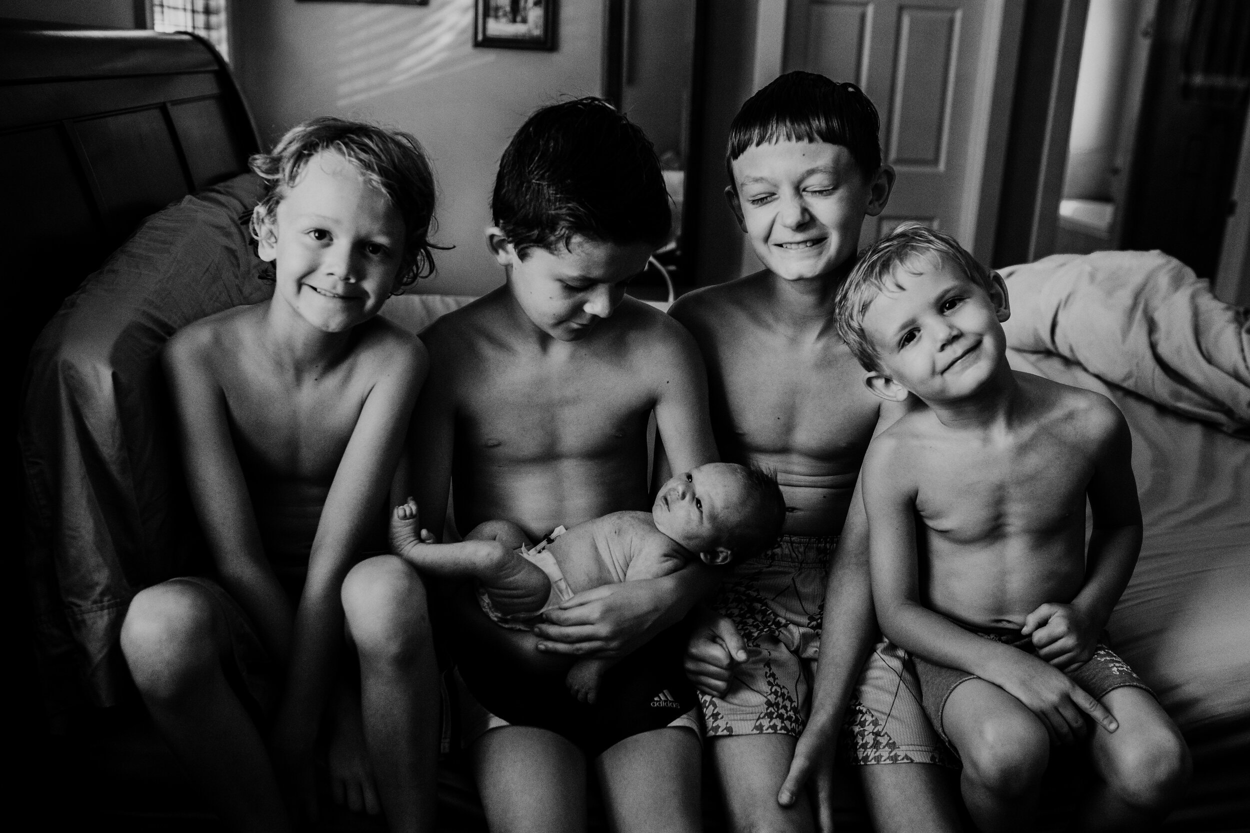 four brothers sit on bed laughing while holding their newborn brother 