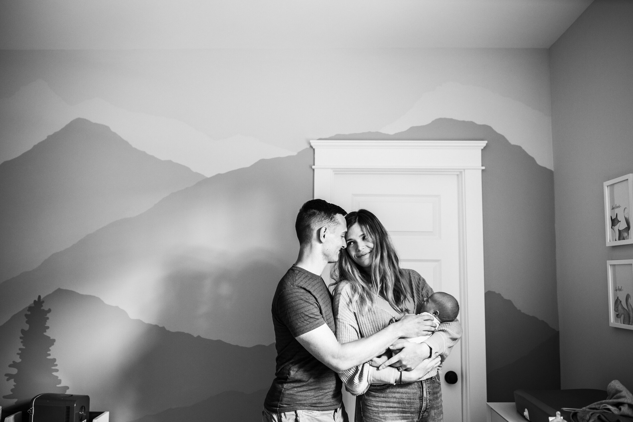 couple stand together holding newborn in nursery with mountains painted on walls